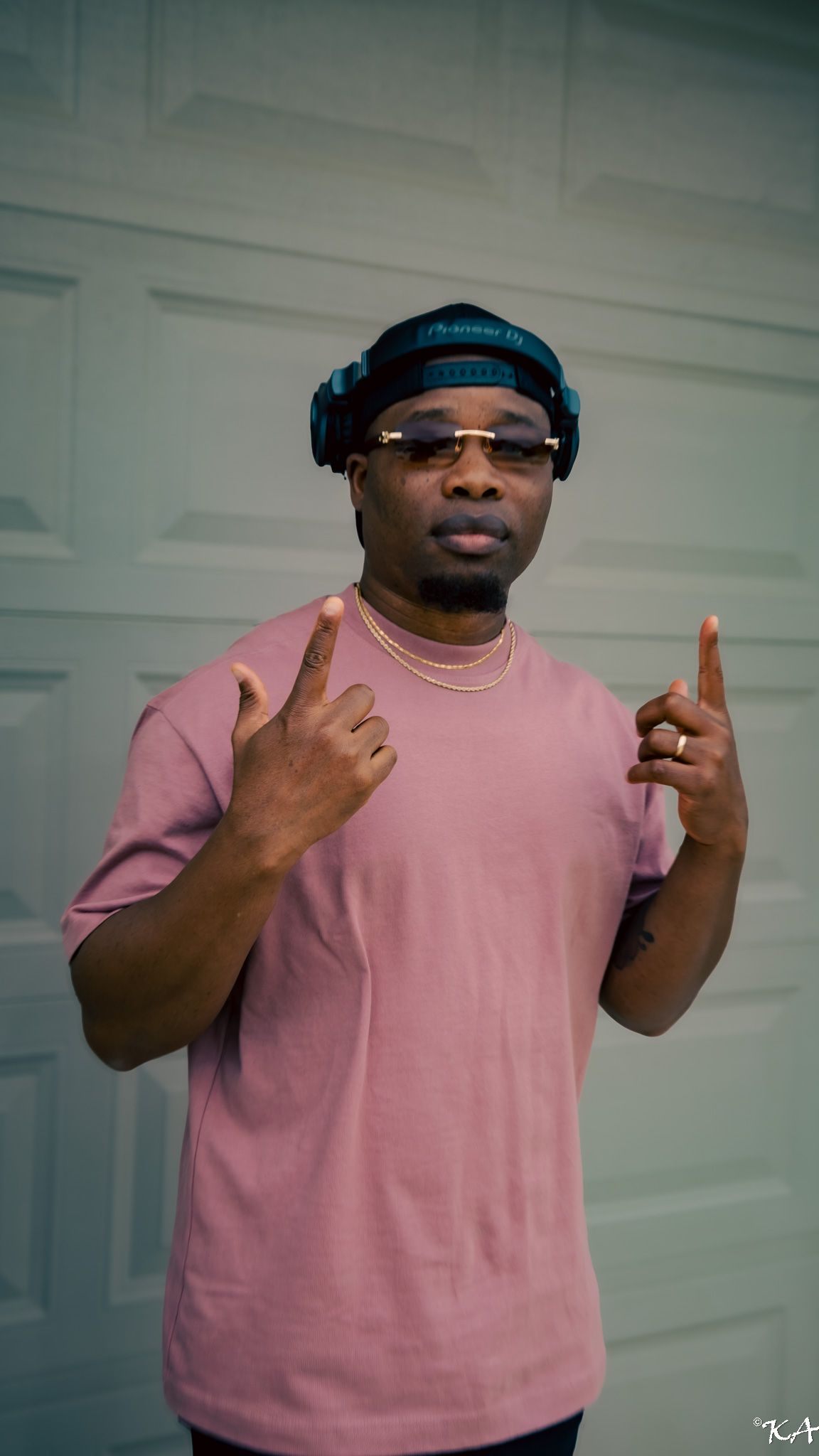 Man in pink t-shirt wearing blue headphones and sunglasses pointing fingers upward near a garage door.