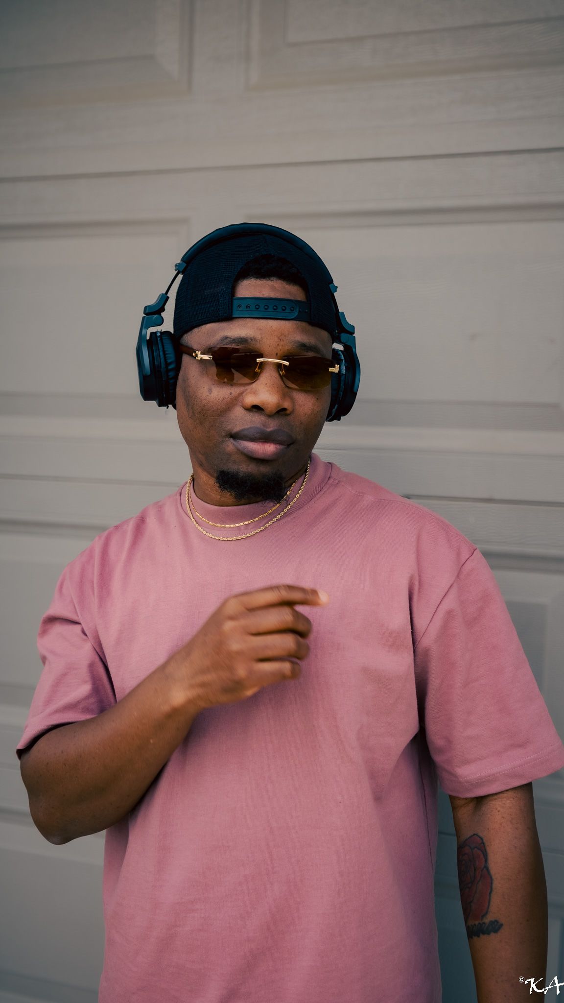 Man in pink t-shirt wearing blue headphones and cap, pointing at camera outdoors.