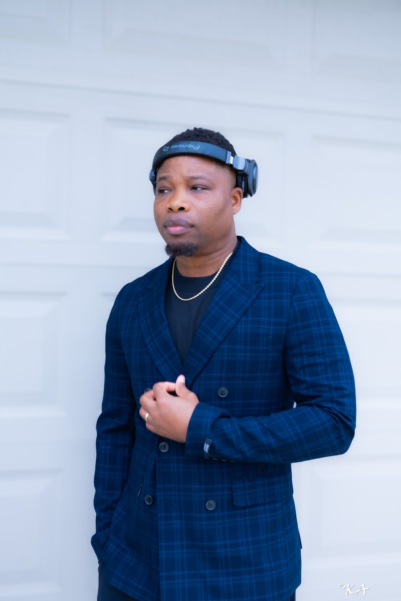 Man in blue plaid blazer wearing headphones as headband, gold chain necklace, posing against white garage door.
