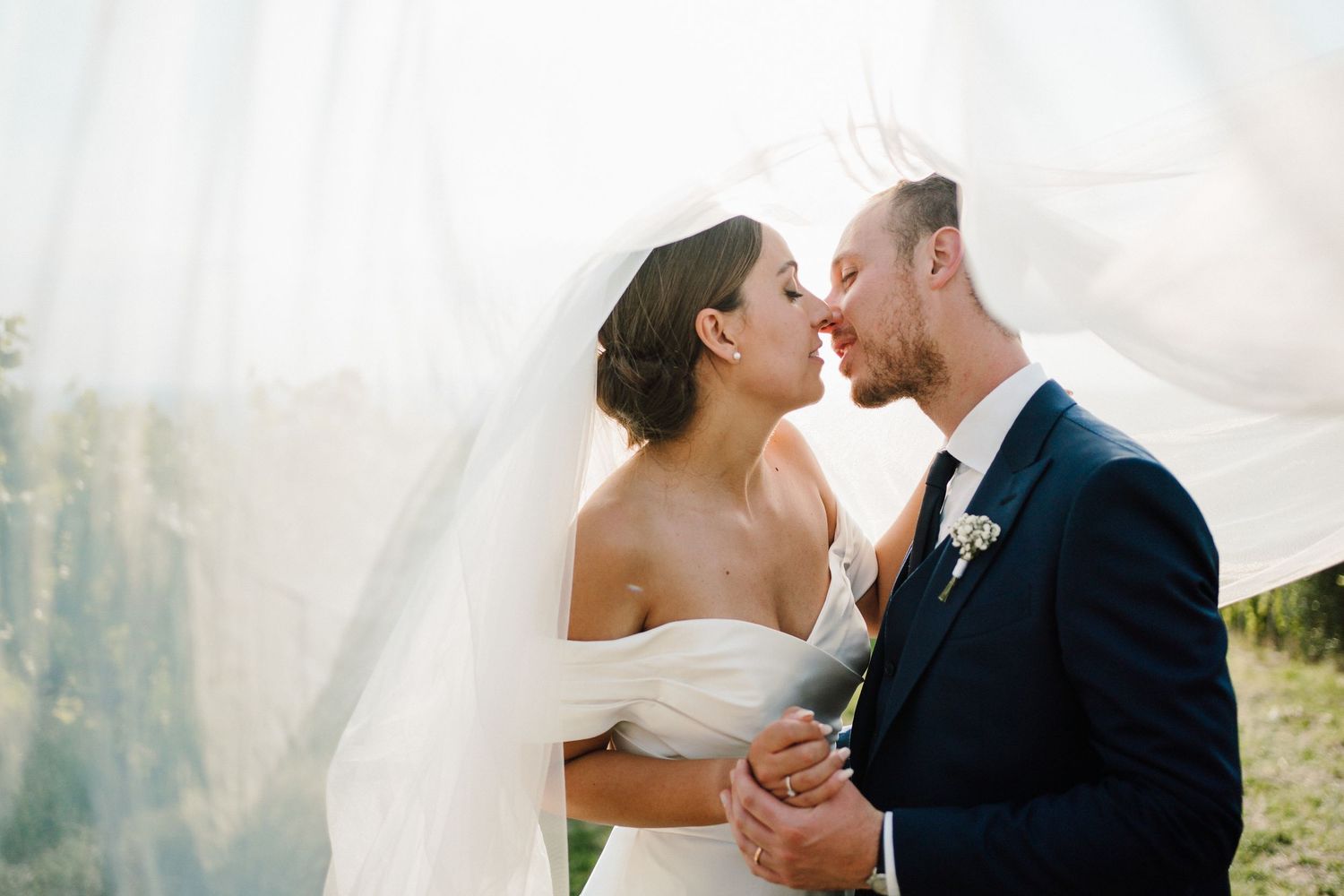 Bride and groom share intimate moment outdoors with flowing veil in sunlight.