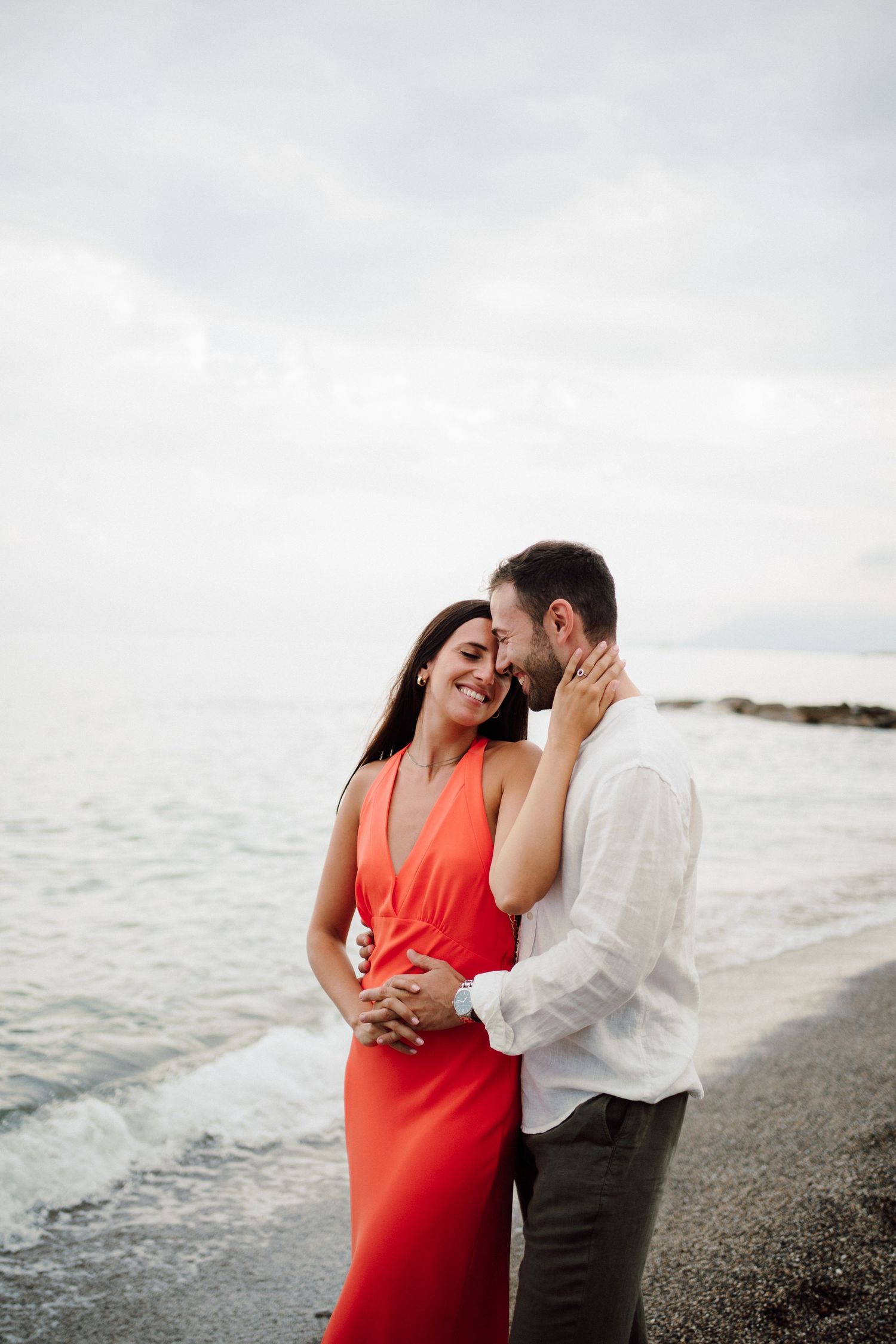 Romantic beachside photoshoot with couple in red dress and white shirt against ocean backdrop.