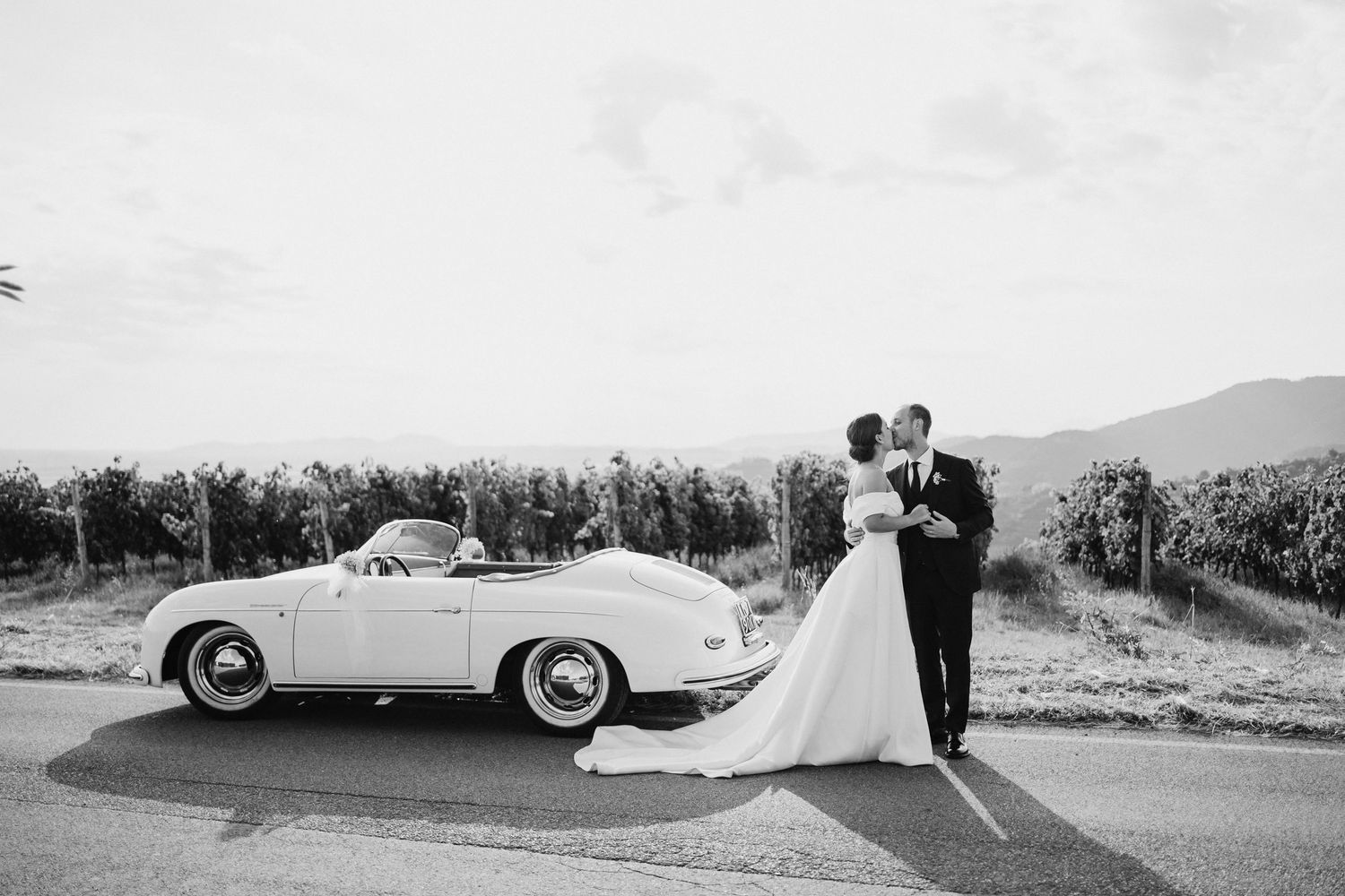 Vintage black and white photograph showing a couple posing with a classic convertible car in a scenic outdoor setting.