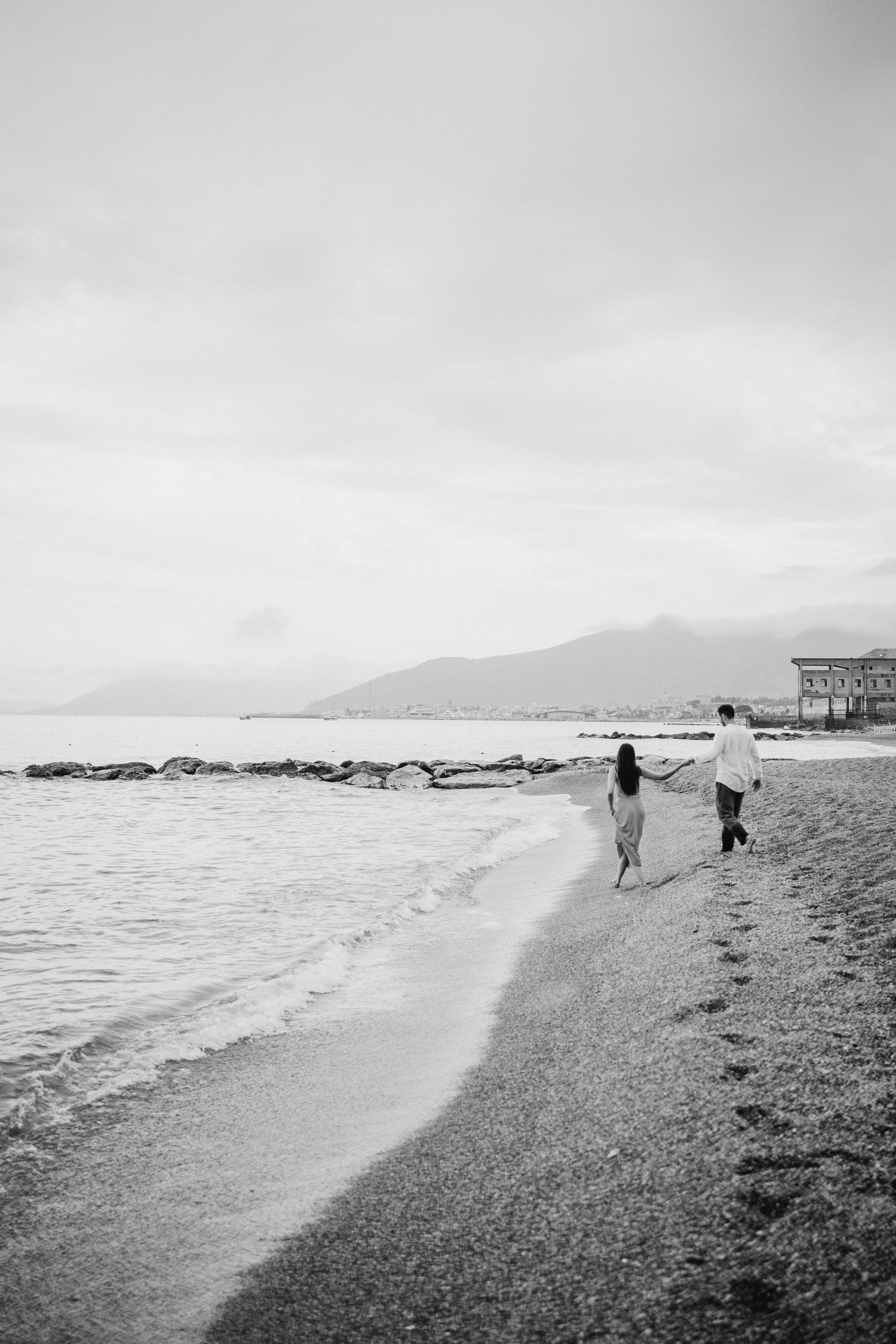 Black and white photograph of a scenic beach shoreline with mountains in the distance.