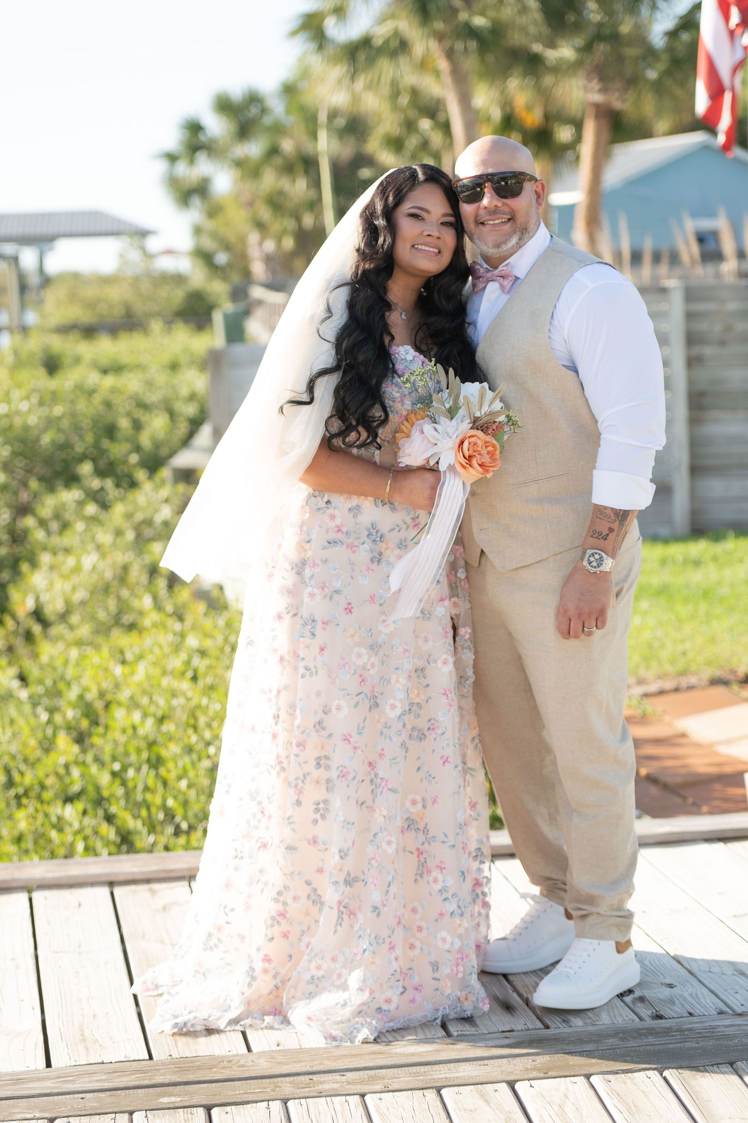 A bride in a floral gown and groom in tan suit pose together on a wooden deck at an outdoor wedding.