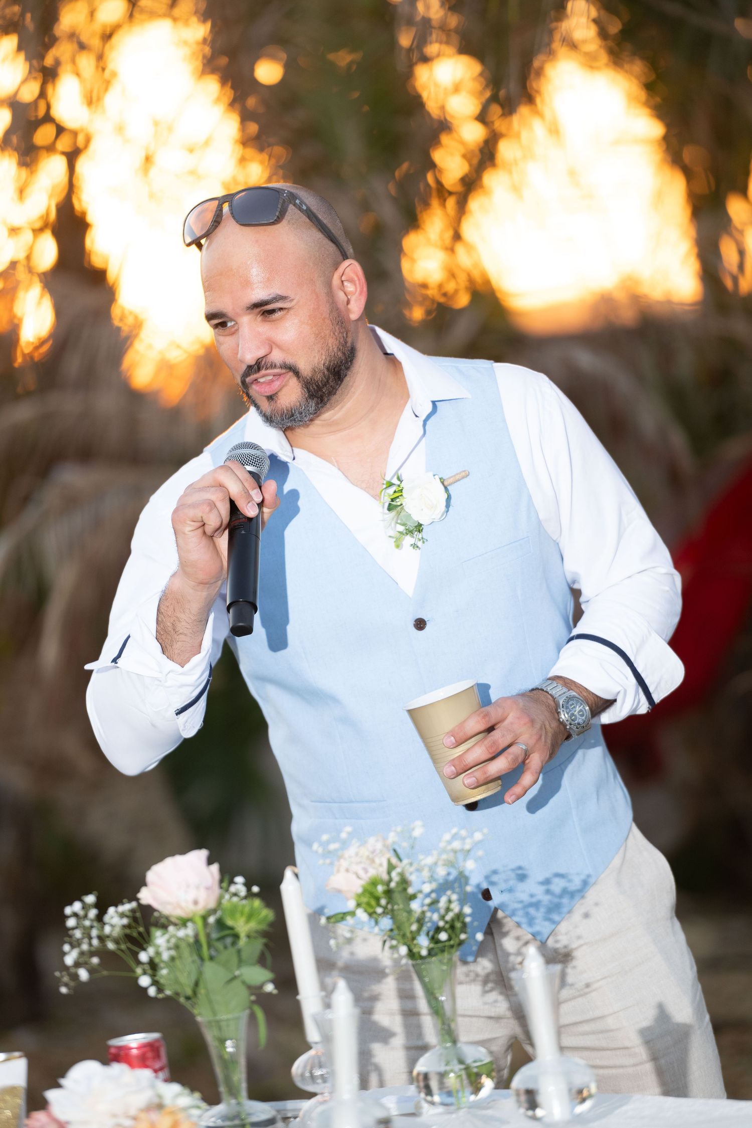 Man in blue vest speaking into microphone at outdoor wedding reception during golden hour sunset.
