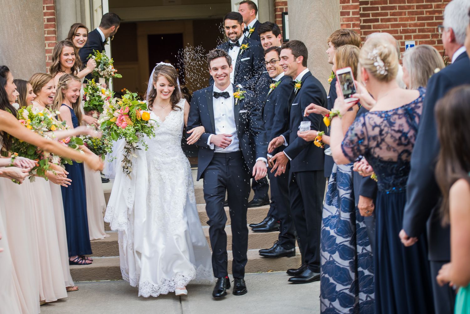 Wedding guests line up outside a brick church building to celebrate the newlyweds' exit with bubbles and cheers.