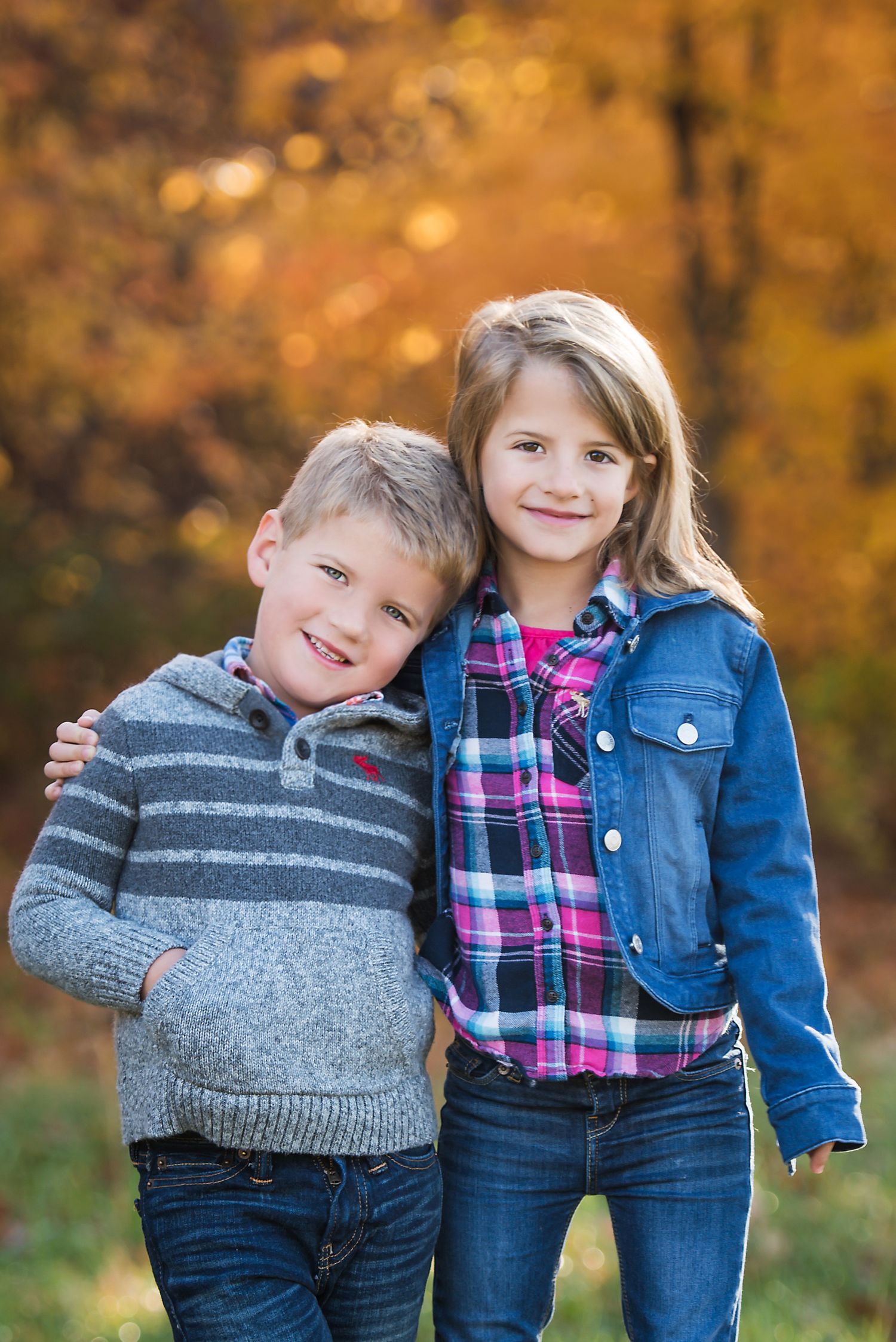 Two young children in casual fall clothing pose together against a backdrop of autumn foliage with golden colors.
