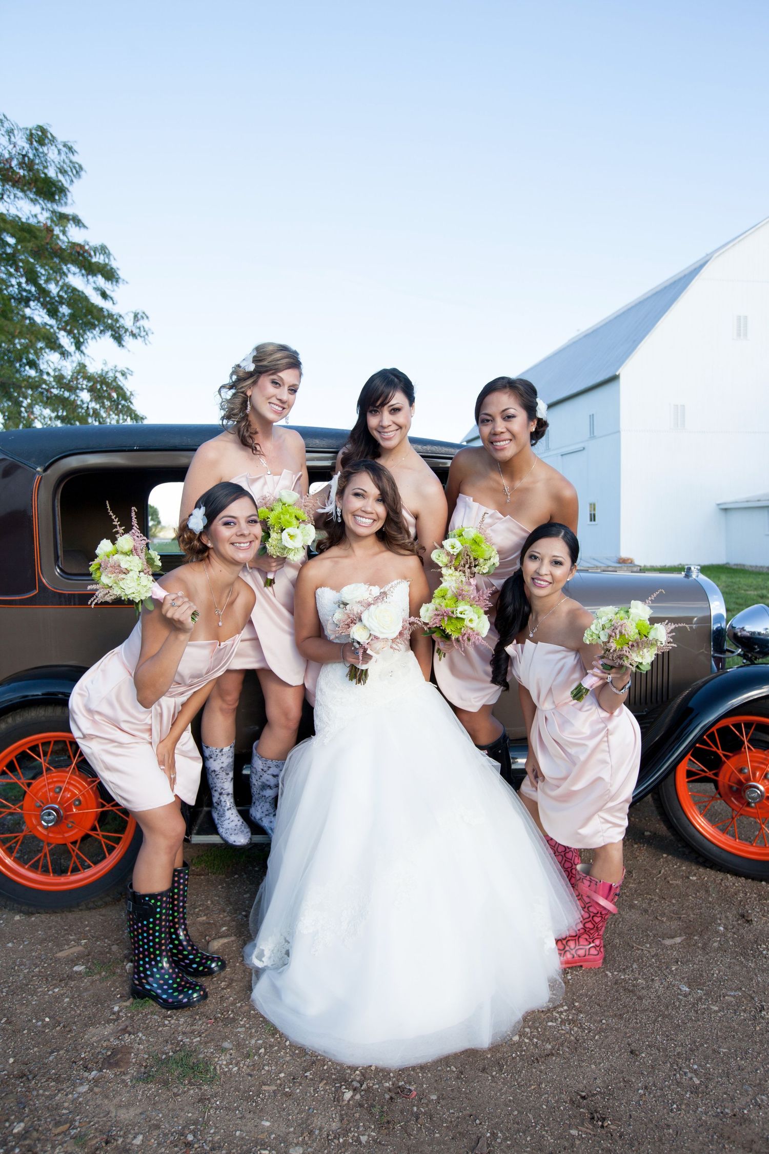 Bridal party in pink dresses and boots posing playfully next to vintage car with red wheels.