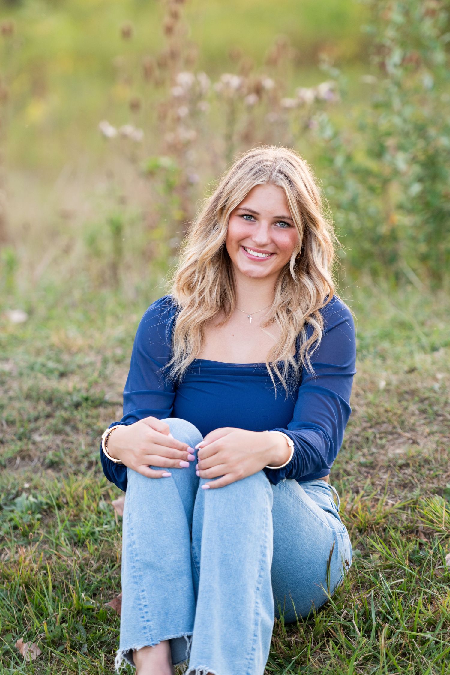 Person wearing blue sweater and light jeans sits casually on grass in outdoor portrait session.