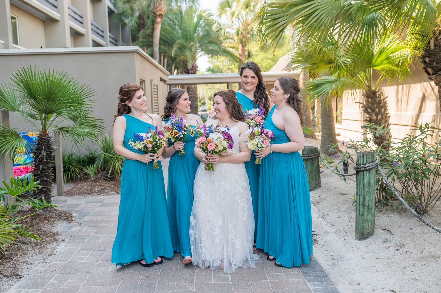 Wedding party in turquoise bridesmaid dresses poses with bride on beach boardwalk near palm trees. Hilton Head Tiki beach wedding