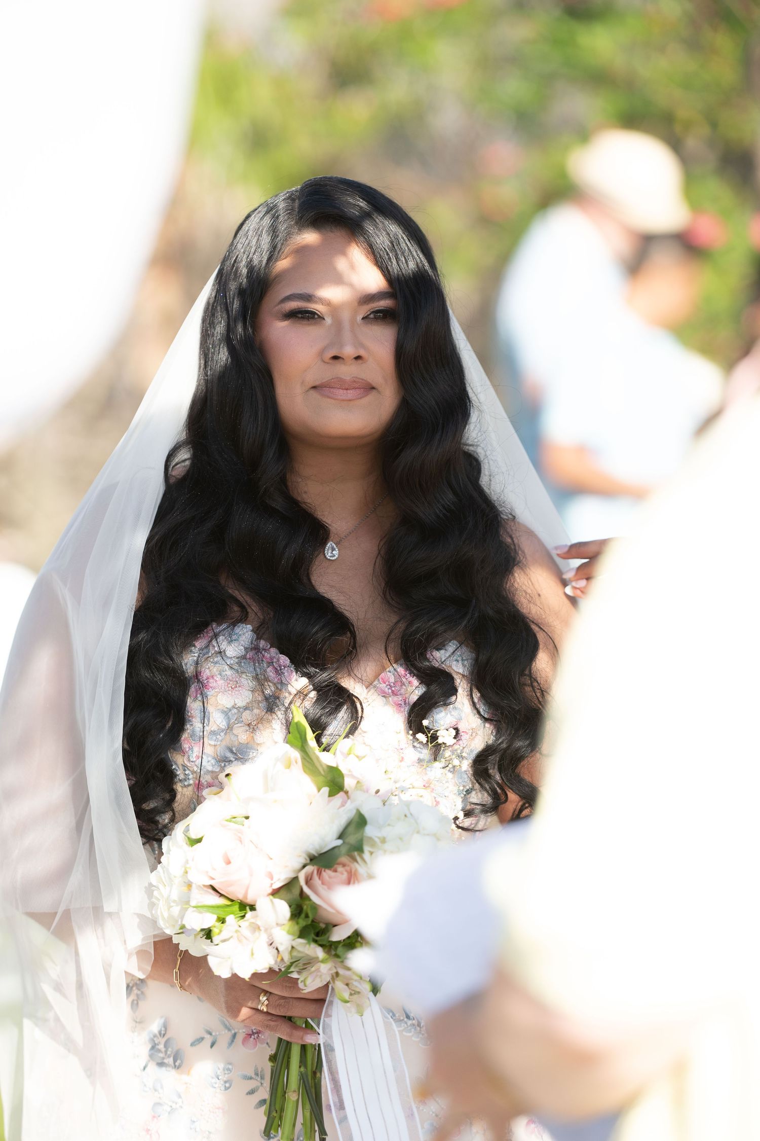 Bride with long wavy black hair wearing a floral gown and veil, holding a white bouquet at an outdoor wedding.
