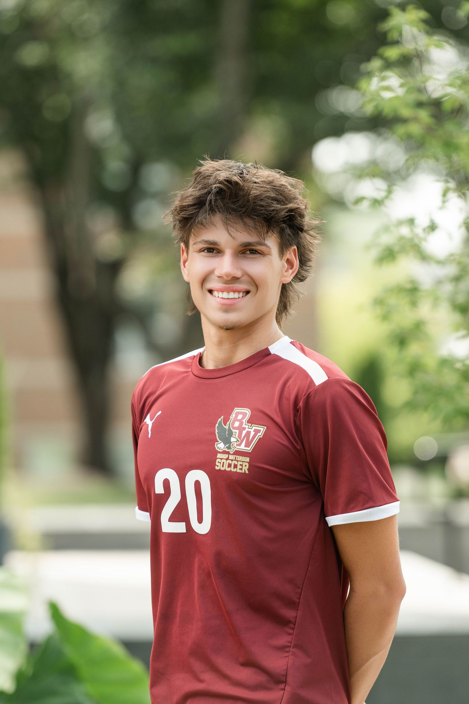 Soccer player wearing maroon jersey number 20 poses outdoors with trees in background.