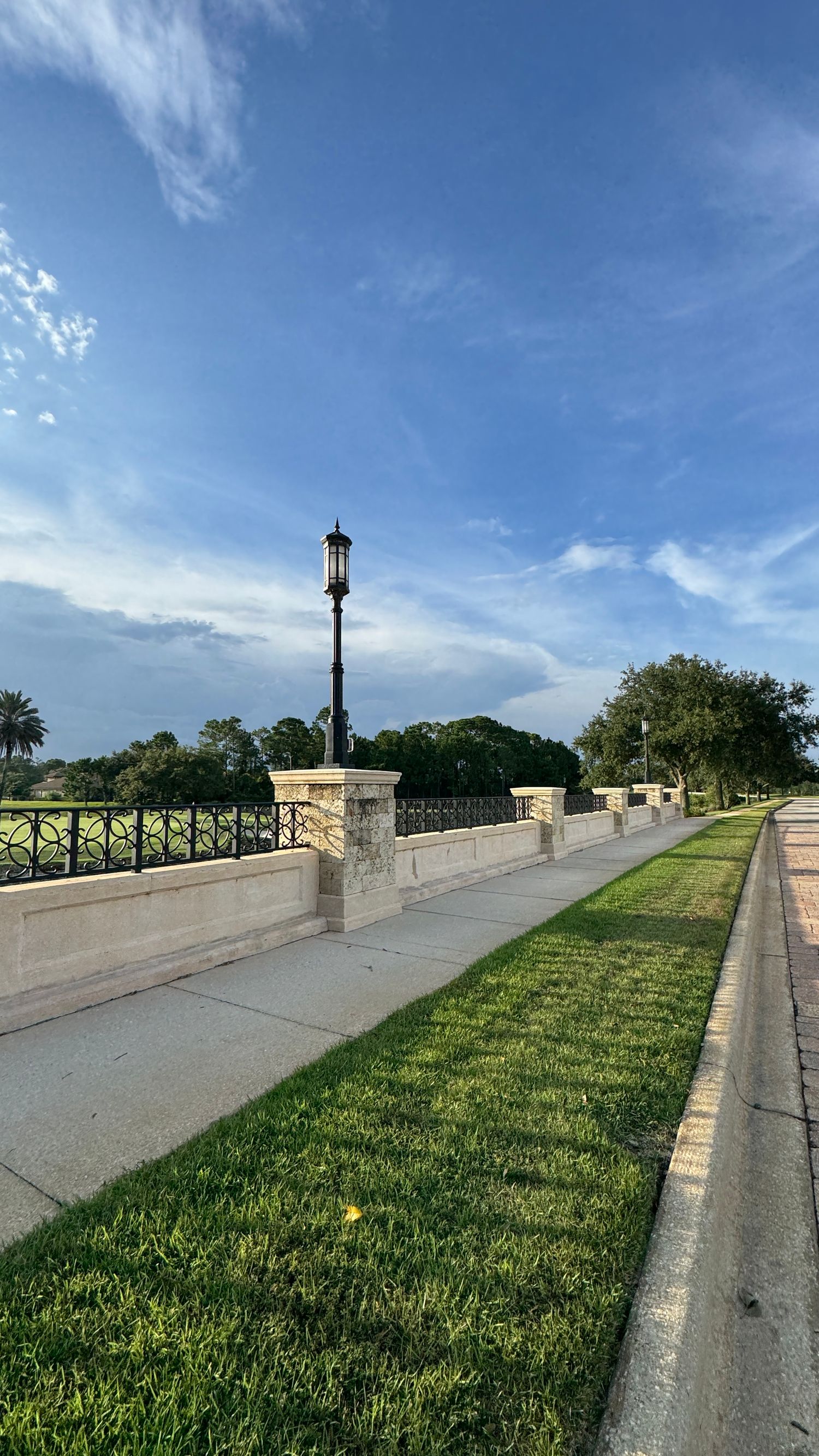 A scenic walkway with ornate iron fencing, stone pillars, and a classic lamp post under a bright blue sky with wispy clouds.