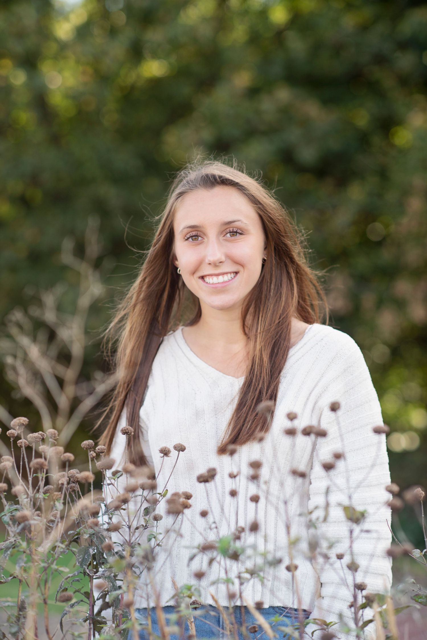 A person in a white sweater sits among dried wildflowers in a natural outdoor setting.