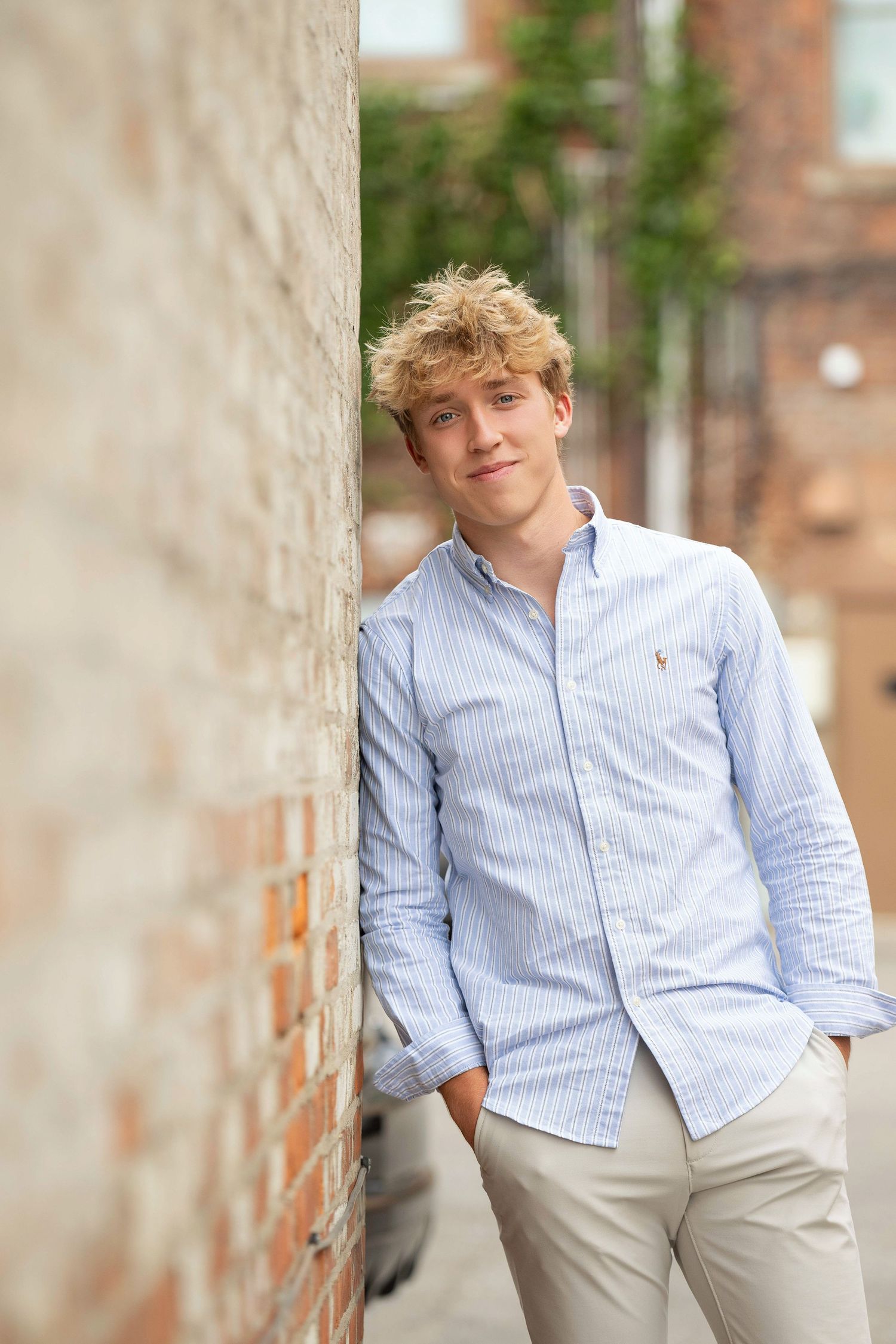 Young person in light blue button-down shirt and khaki pants poses casually against stone wall in outdoor setting.