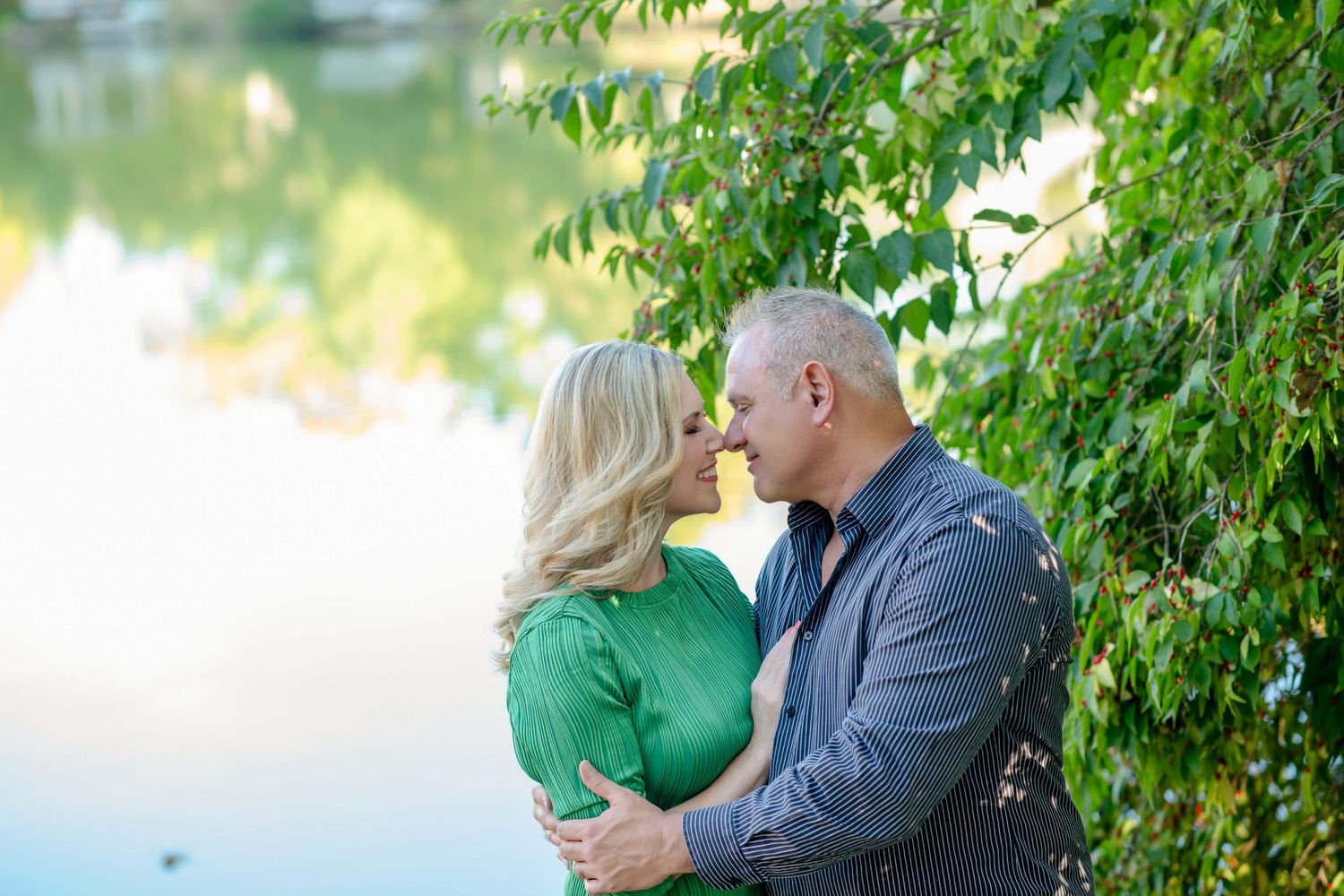 Romantic couple embracing under green trees near water during golden hour.