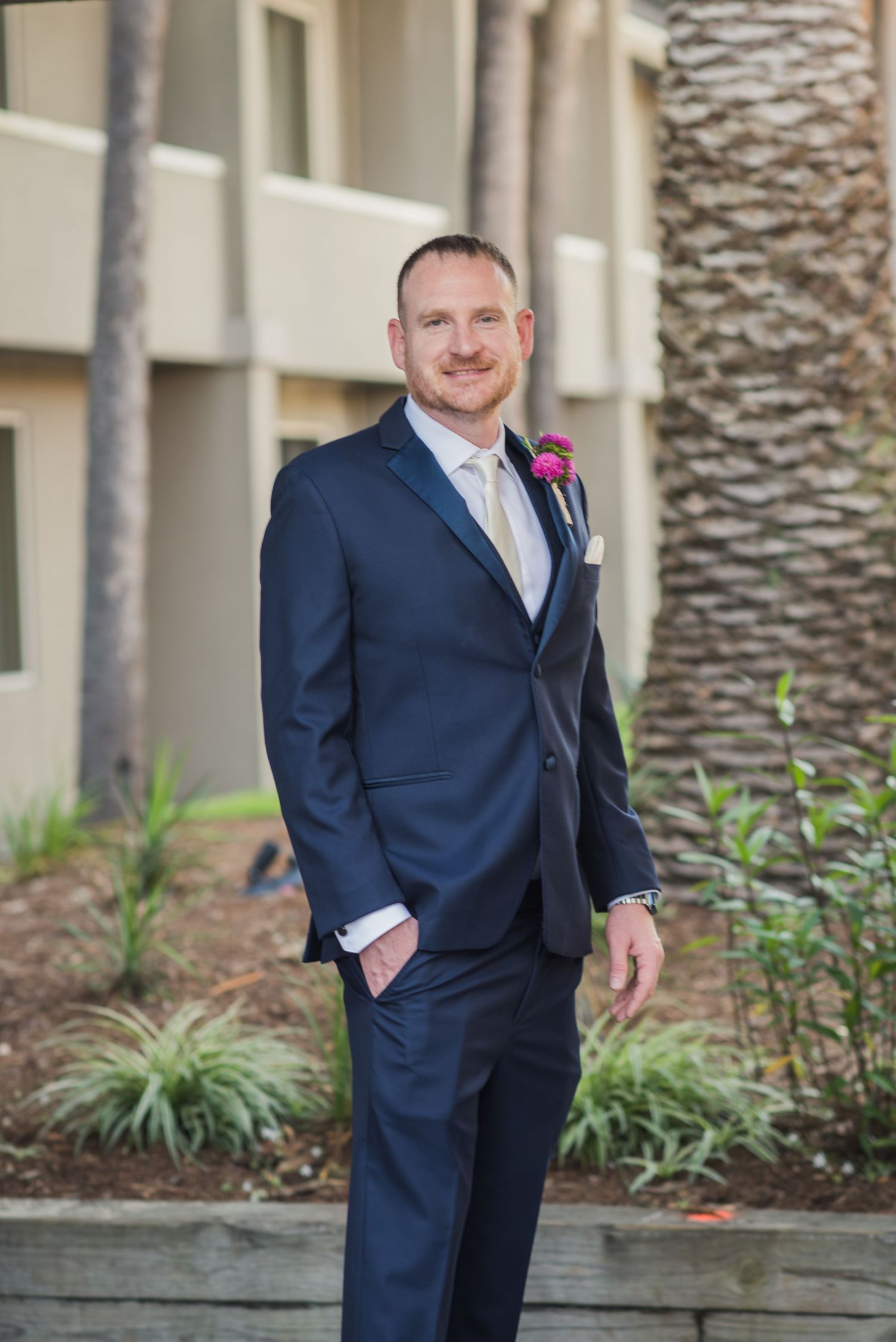 Groomsmen in navy suits stand casually against a building exterior with palm trees.