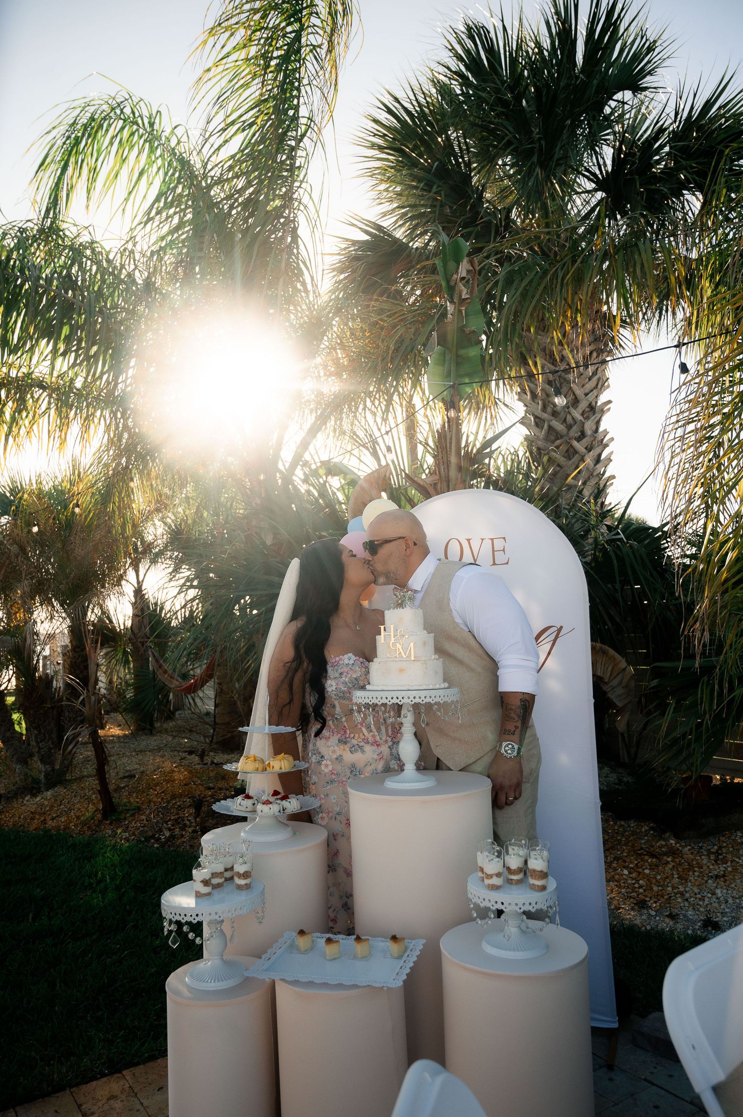 Couple kissing at outdoor wedding cake table with white arch, palm trees, and golden sunset in background.