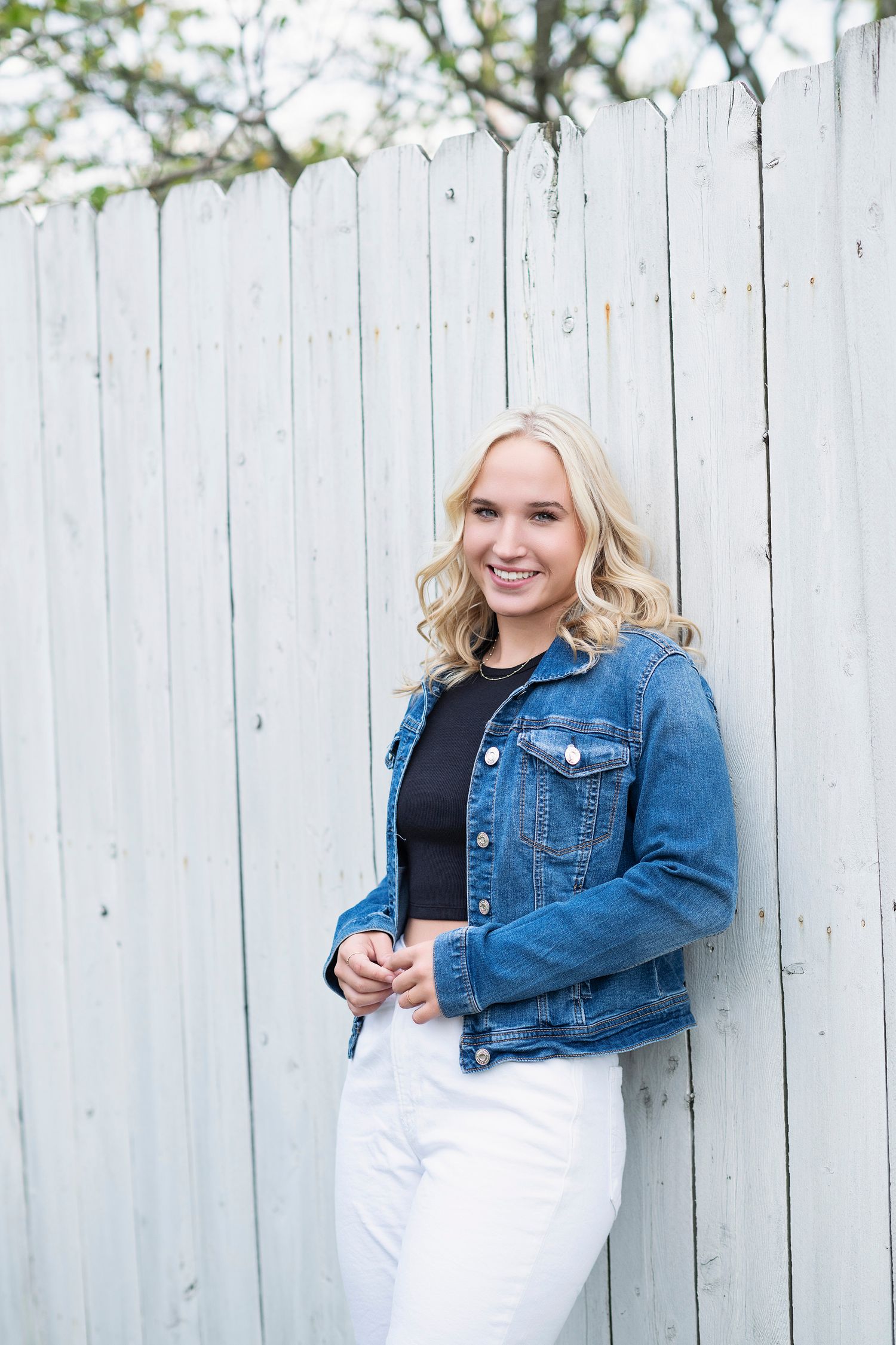 A casual outdoor portrait against a white fence featuring a denim jacket and white pants outfit.