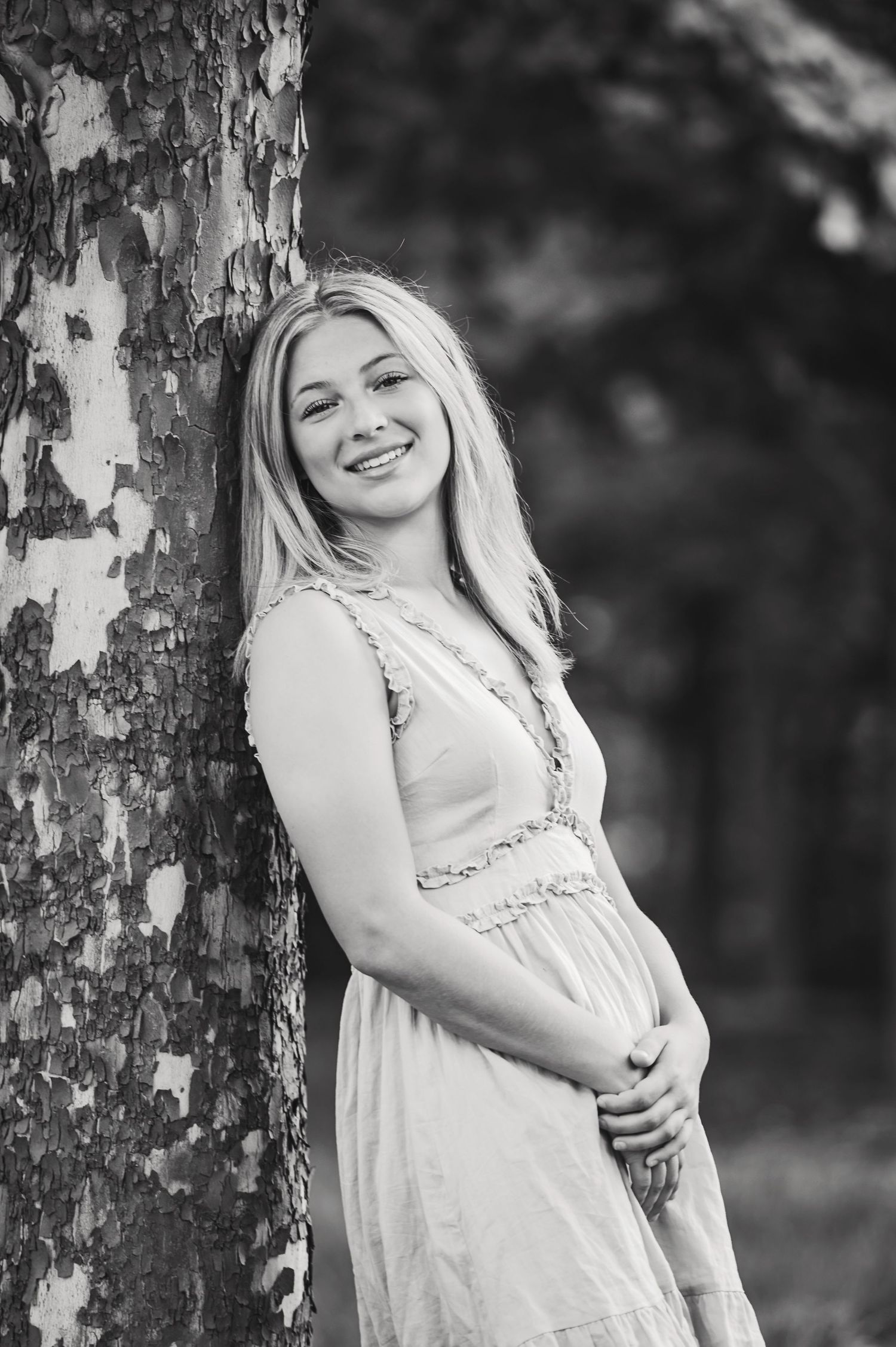 A Black and white senior portrait in a white dress poses against a birch tree trunk in a black and white artistic portrait series.