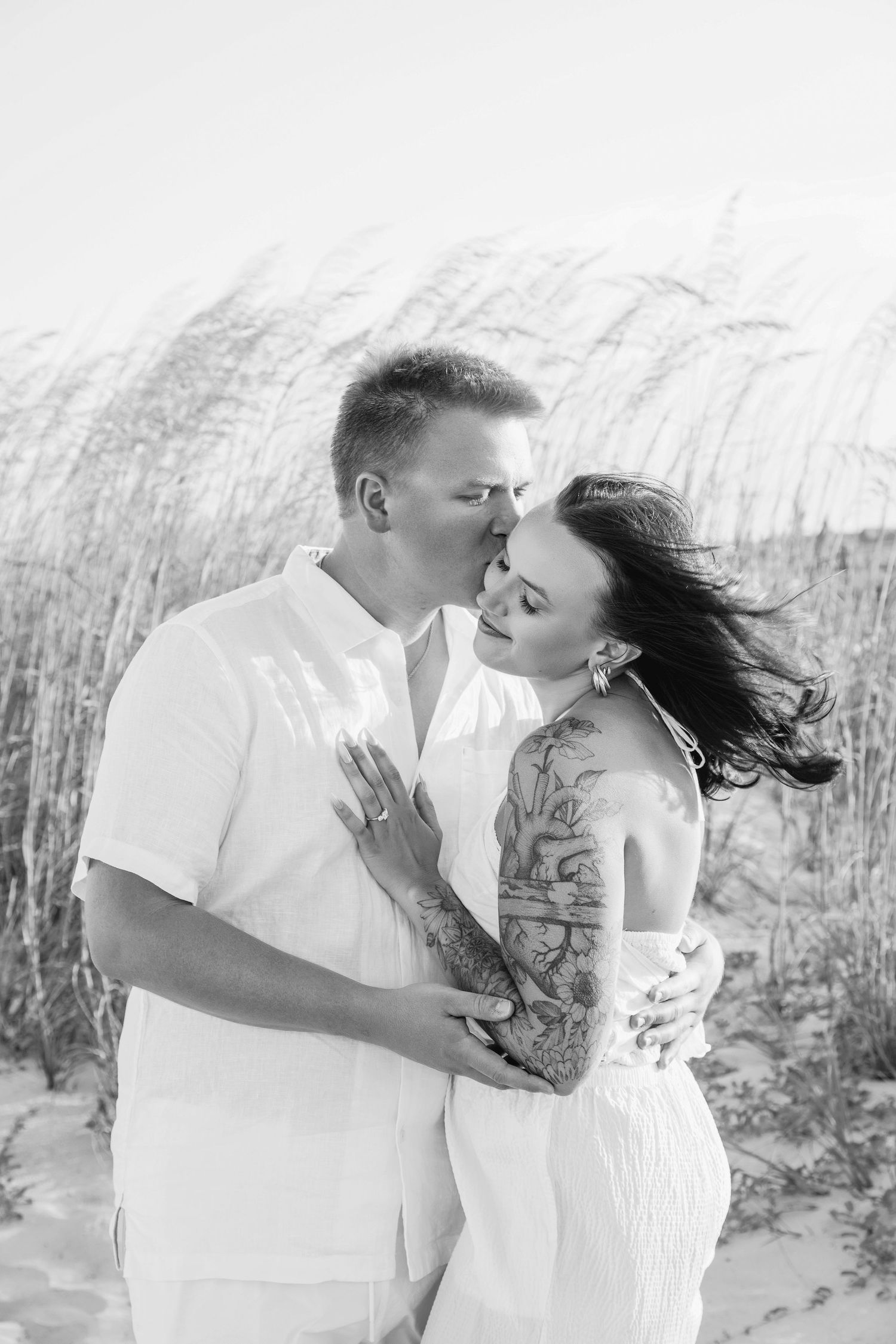 Black and white photo of a couple embracing on a sandy beach, woman with tattooed arm in white dress, man in white shirt.