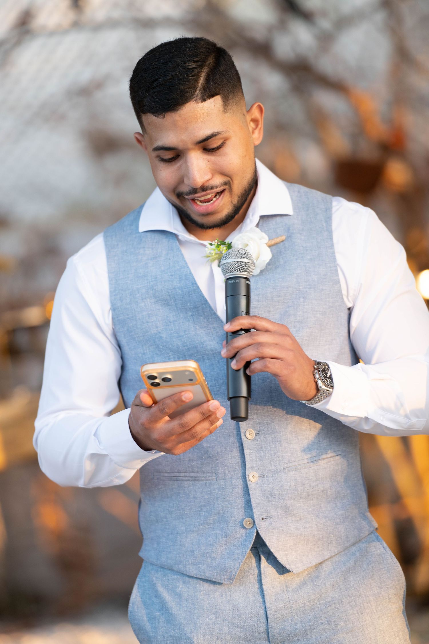 Smiling groom in light blue vest holds microphone and phone, reading wedding speech outdoors.