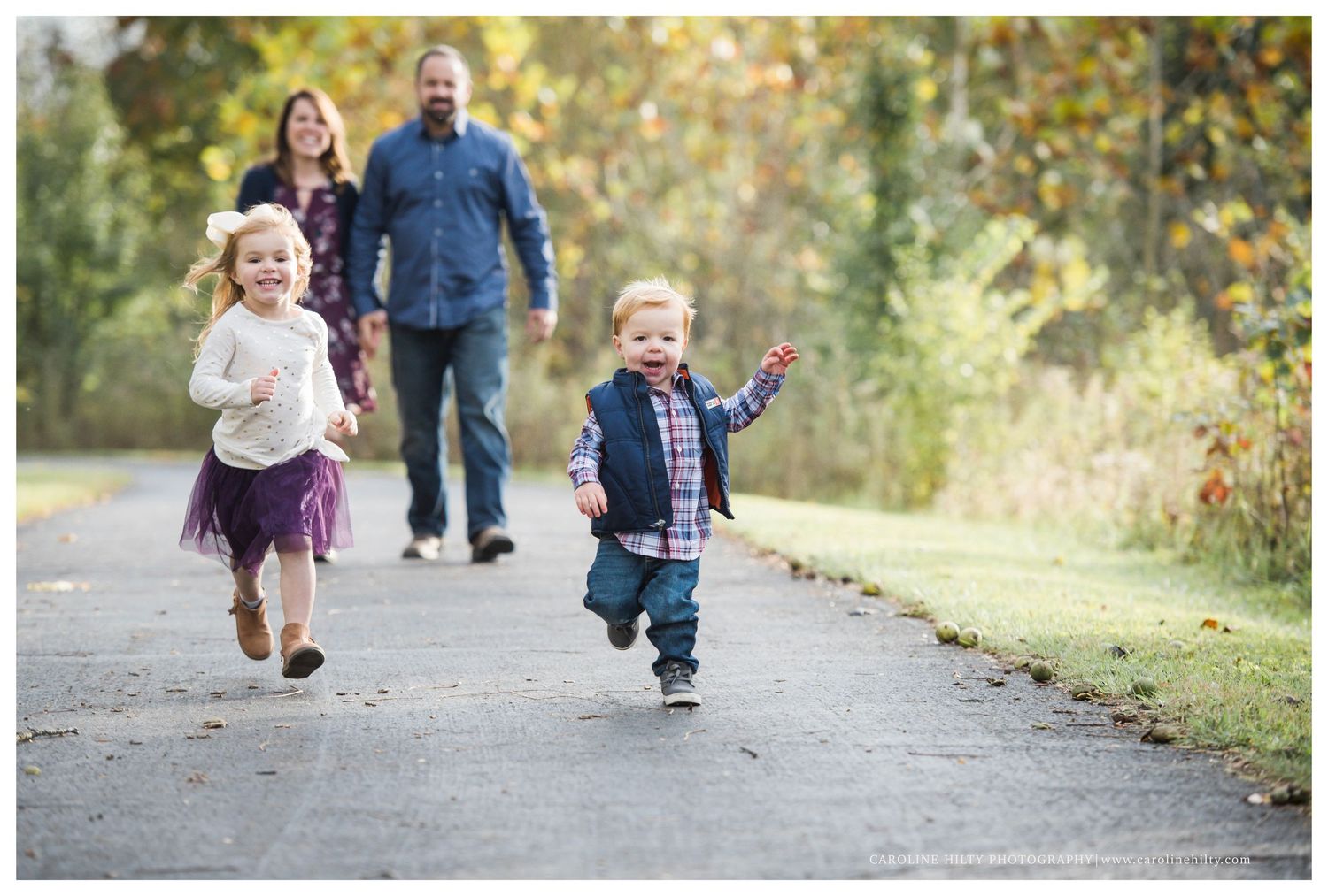 Children running joyfully down a path while adults follow behind on an autumn day.