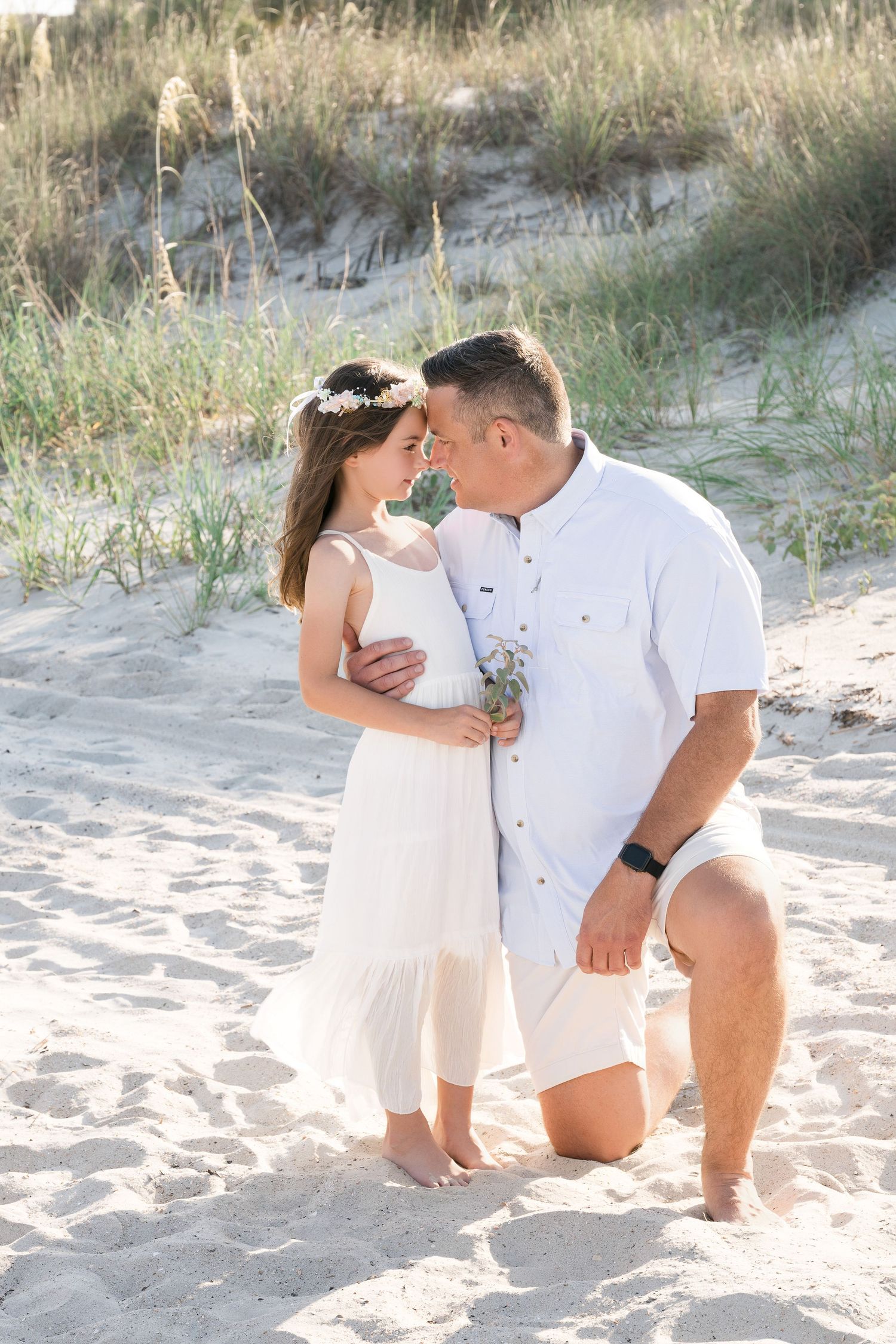 Couple in white attire sharing a romantic moment on sandy beach dunes, man kneeling holding flowers, woman wearing floral crown.