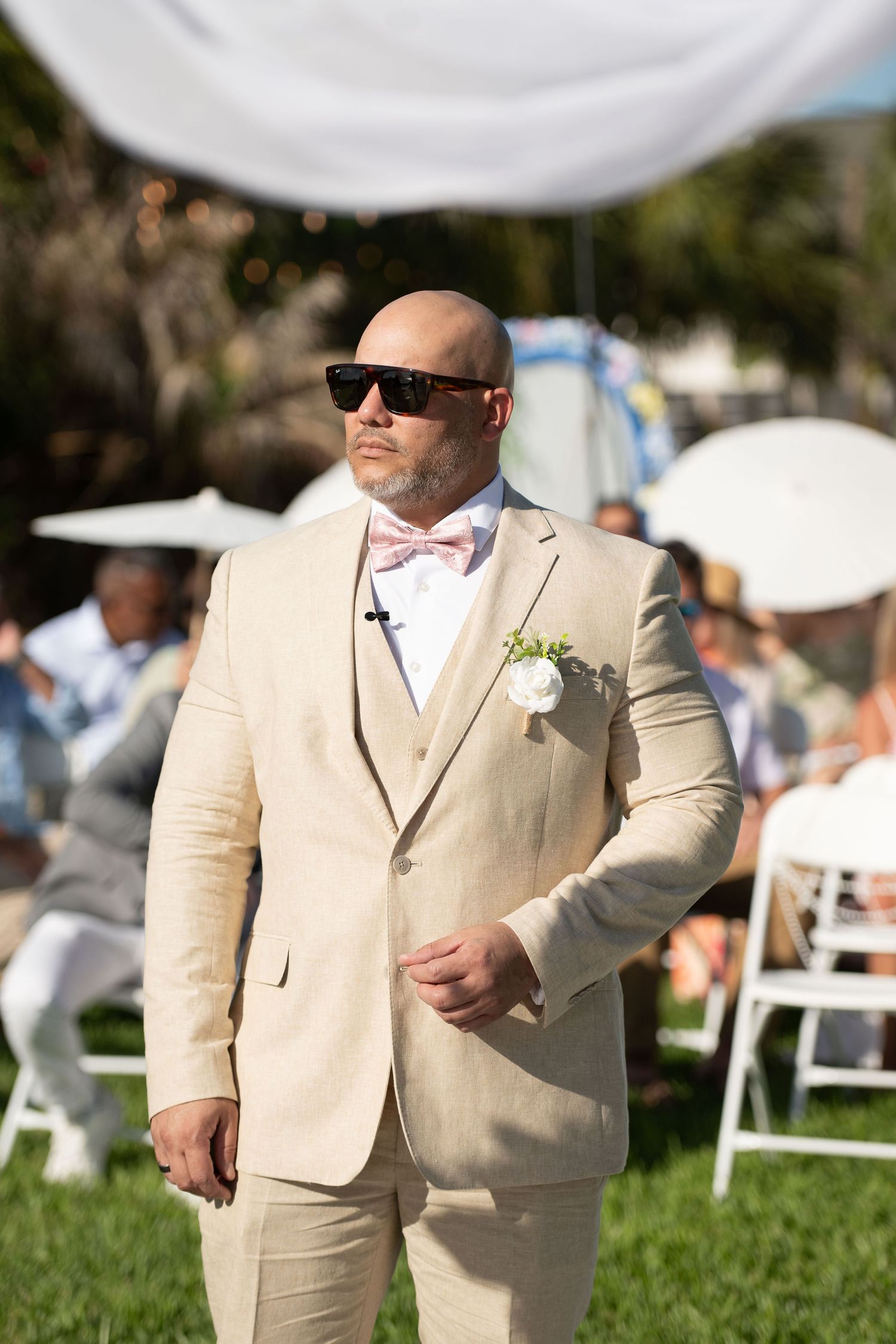Bald groom in beige linen suit, pink bow tie, boutonniere, and sunglasses stands at outdoor wedding ceremony.