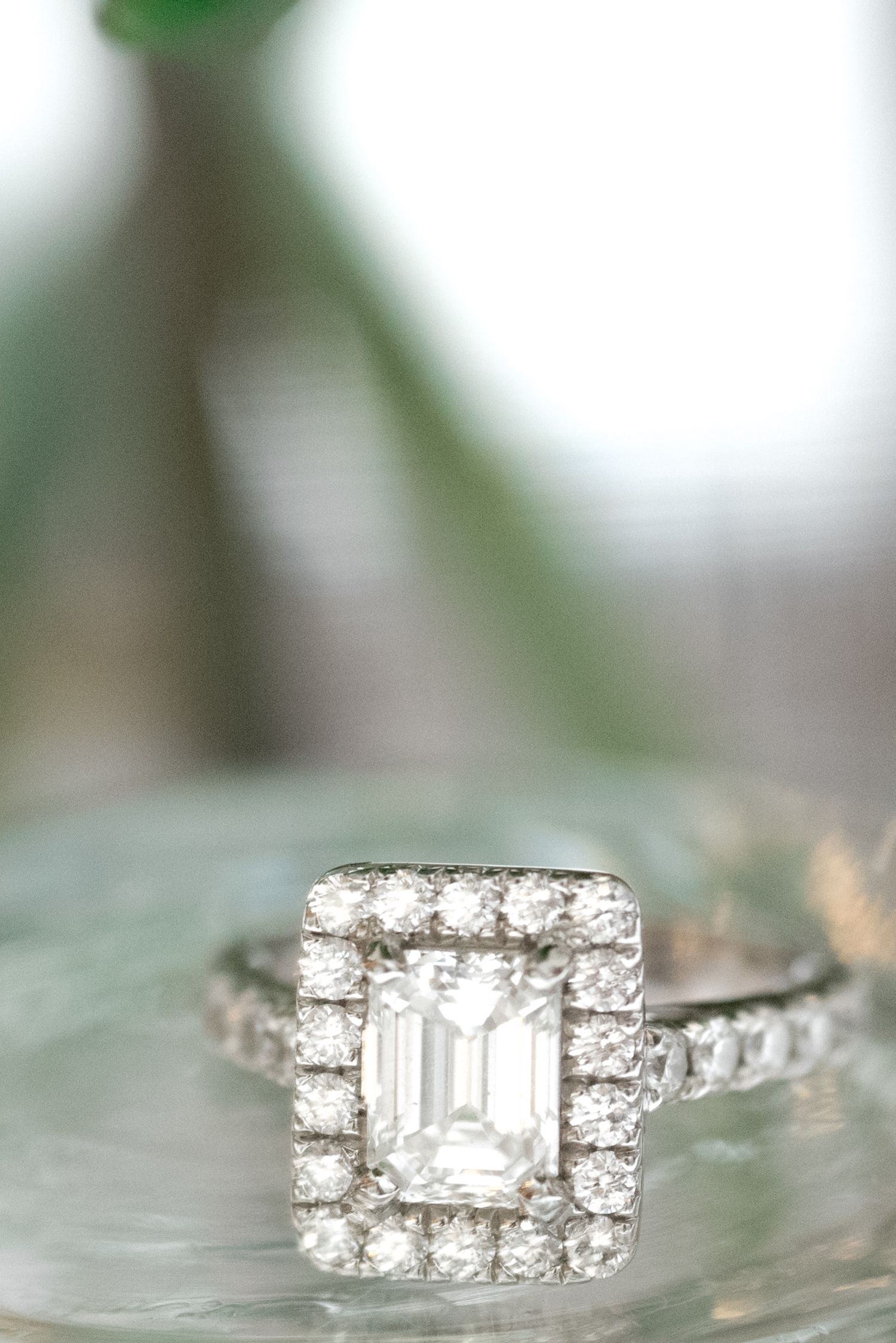 Close-up macro shot of a diamond engagement ring with an emerald cut center stone and halo setting on reflective surface.