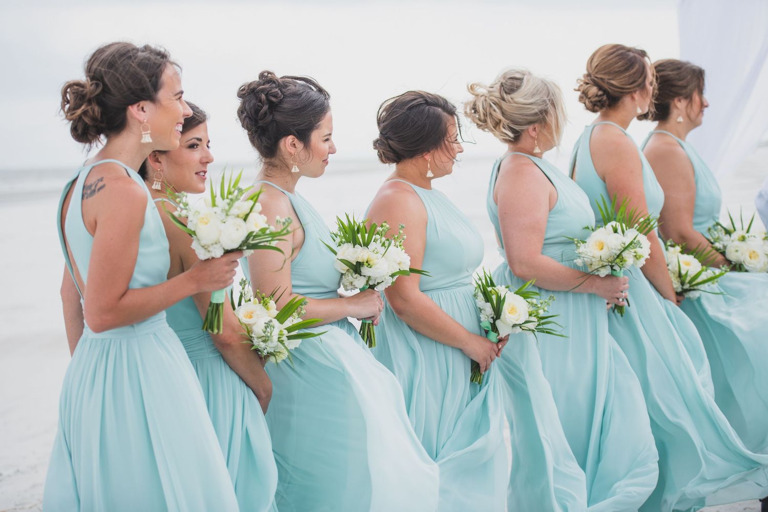 Hilton Head Bridesmaids in matching mint green dresses hold white floral bouquets while laughing on a beach.