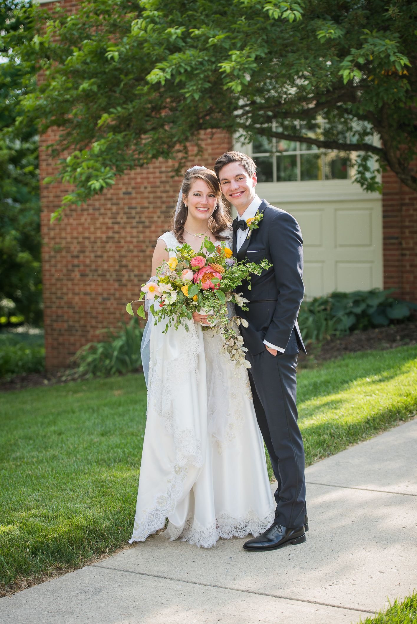 A couple in wedding attire poses together outdoors on a garden path with brick walls and greenery in the background.