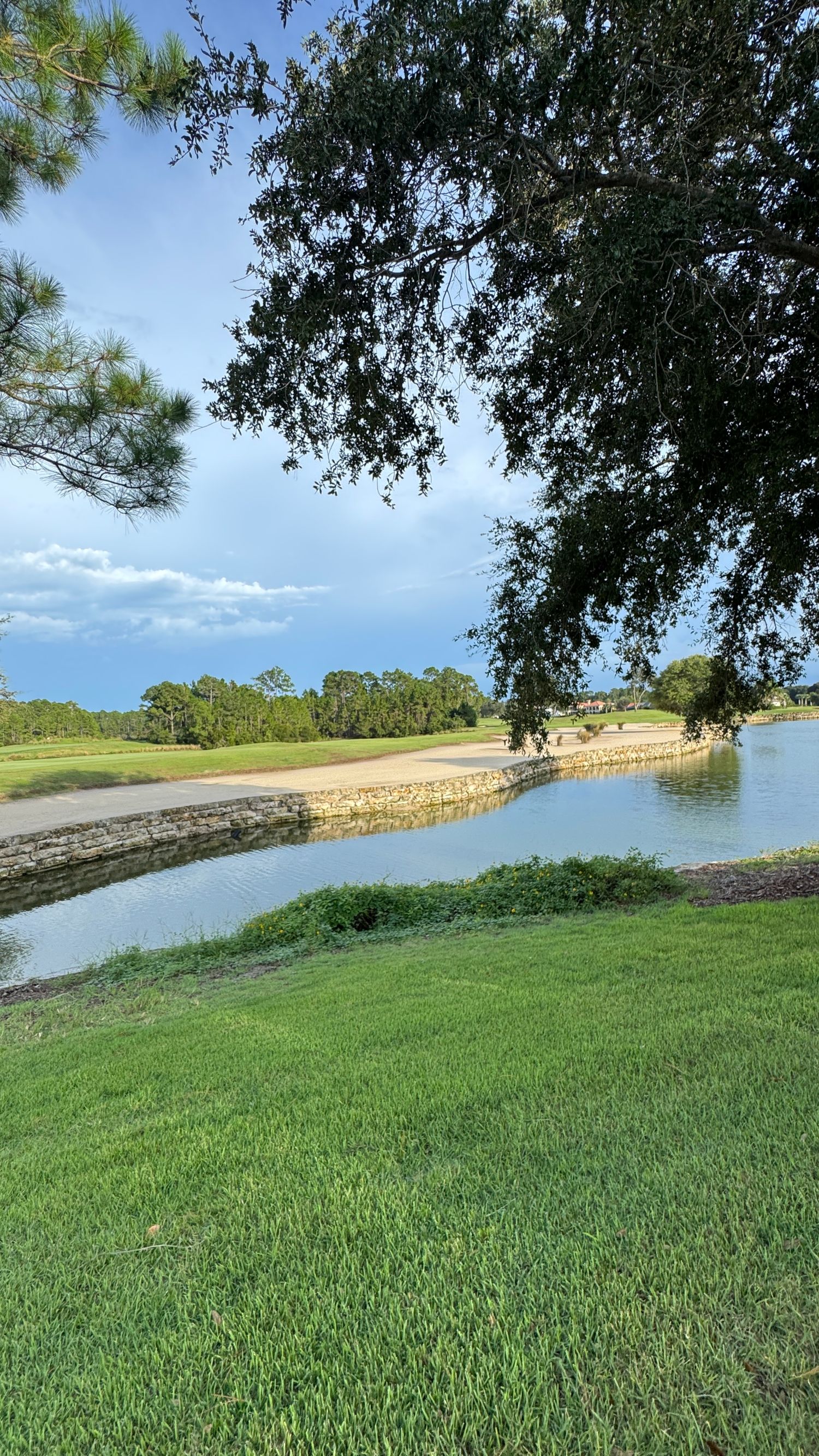 Scenic golf course water hazard with lush green fairways, stone-lined pond, and oak trees under a partly cloudy blue sky.