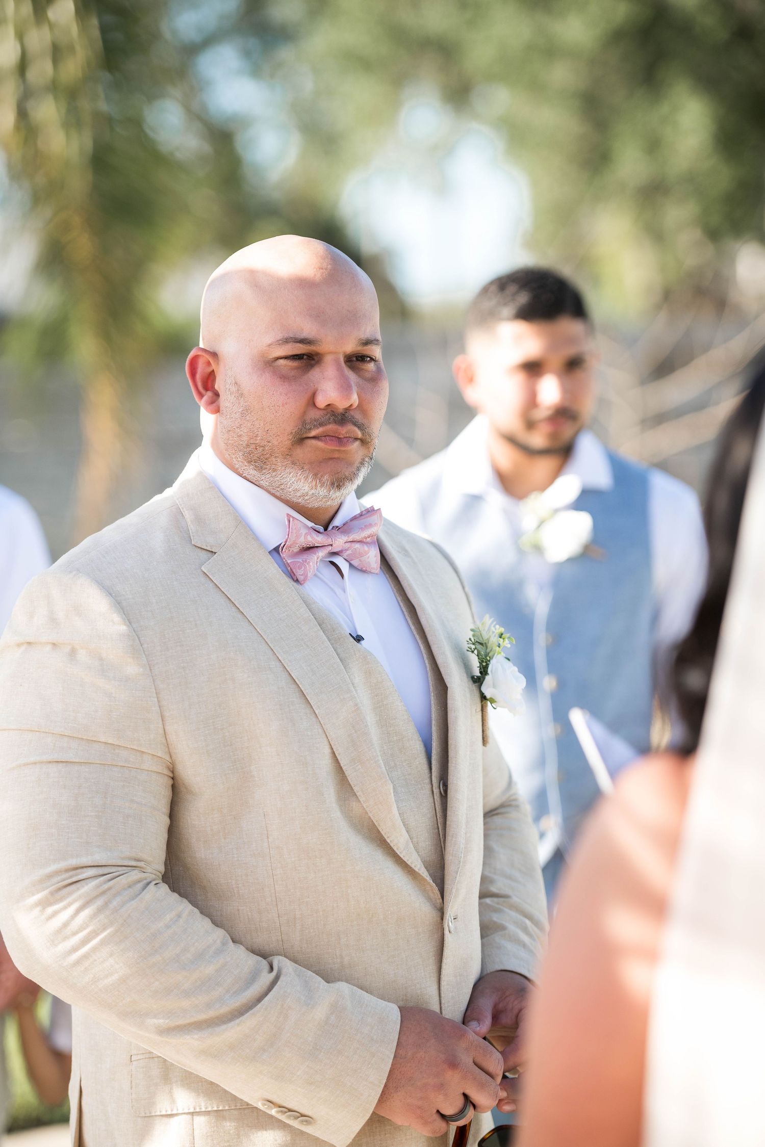 Groom in beige suit with pink bow tie and boutonniere awaits bride at outdoor wedding ceremony.