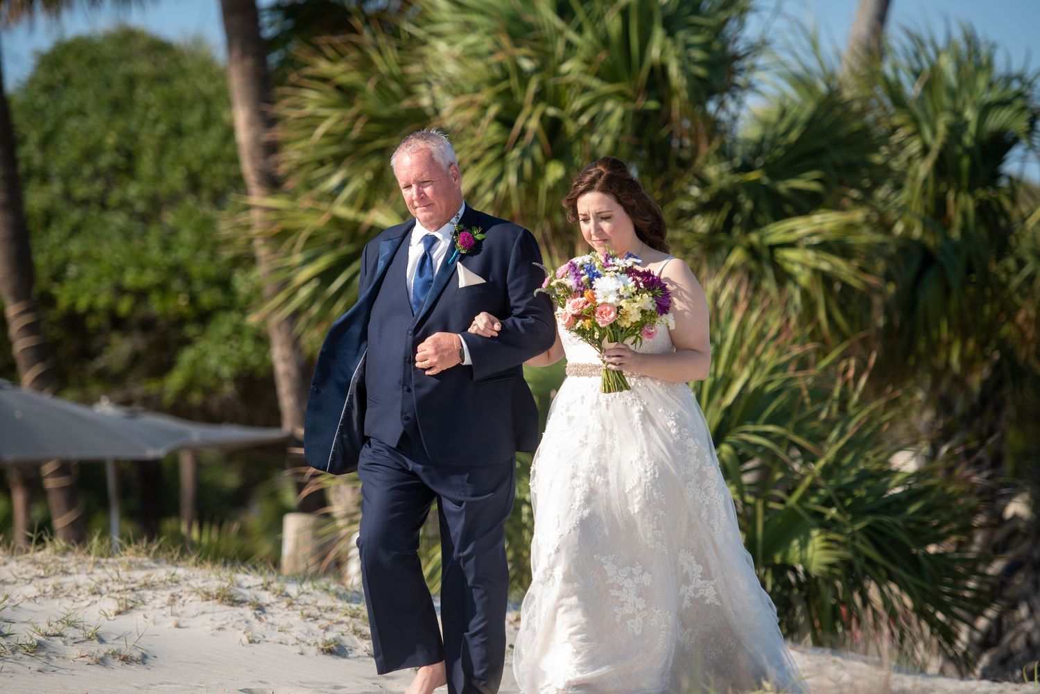 Beach wedding ceremony procession with palm trees and ocean in background.