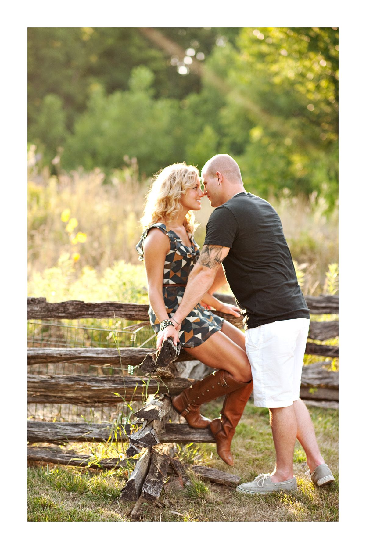 A romantic outdoor engagement photo shoot featuring a couple sitting on rustic wooden fence in a sunlit meadow.