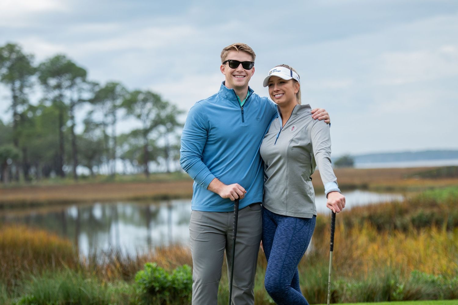 Hilton Head golf couple wearing Southern Tide brand clothing embraces while standing in front of a scenic coastal marsh landscape.