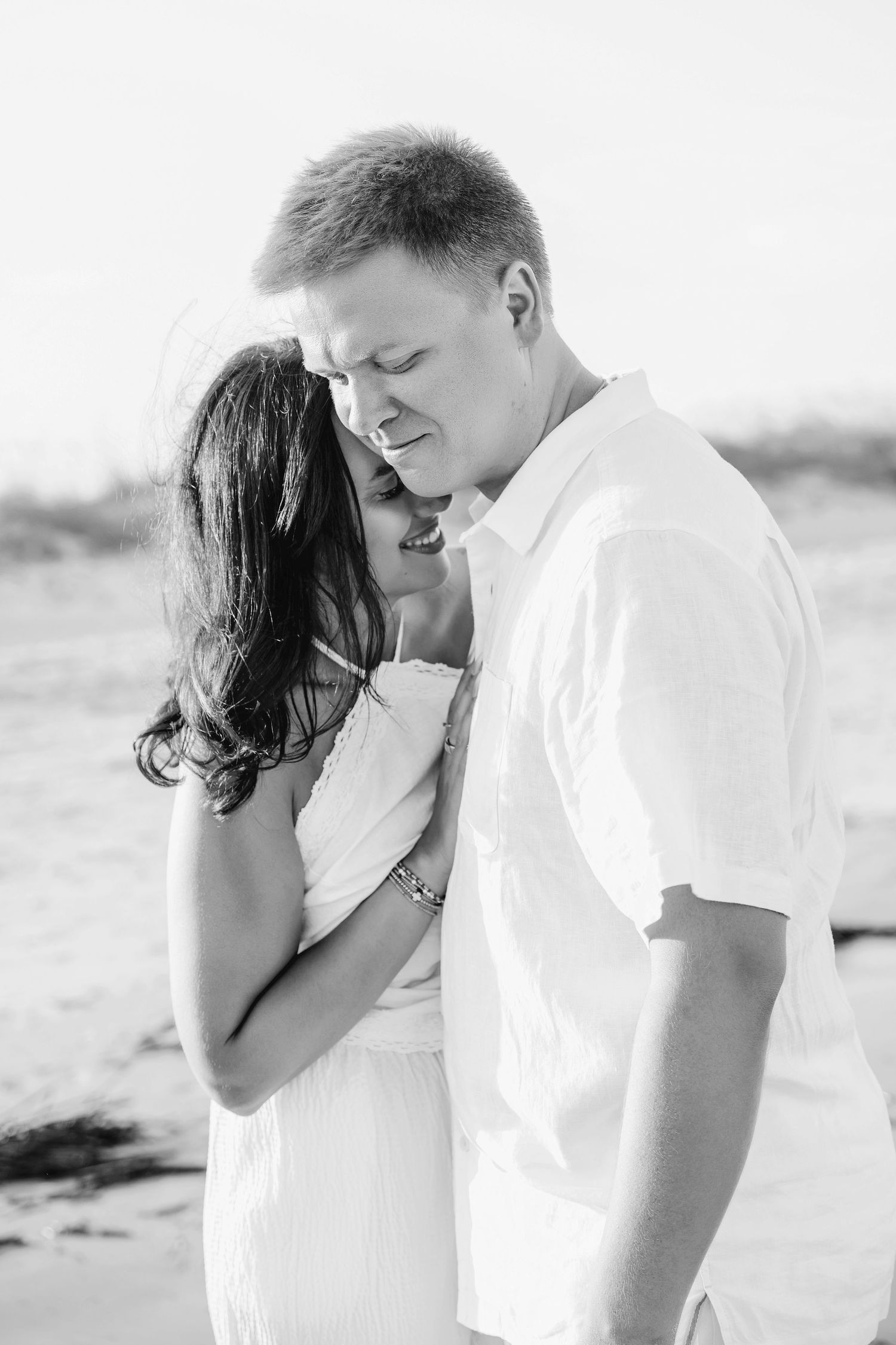 Black and white photo of a couple embracing tenderly on a beach, the man in a white shirt smiling down at the woman.