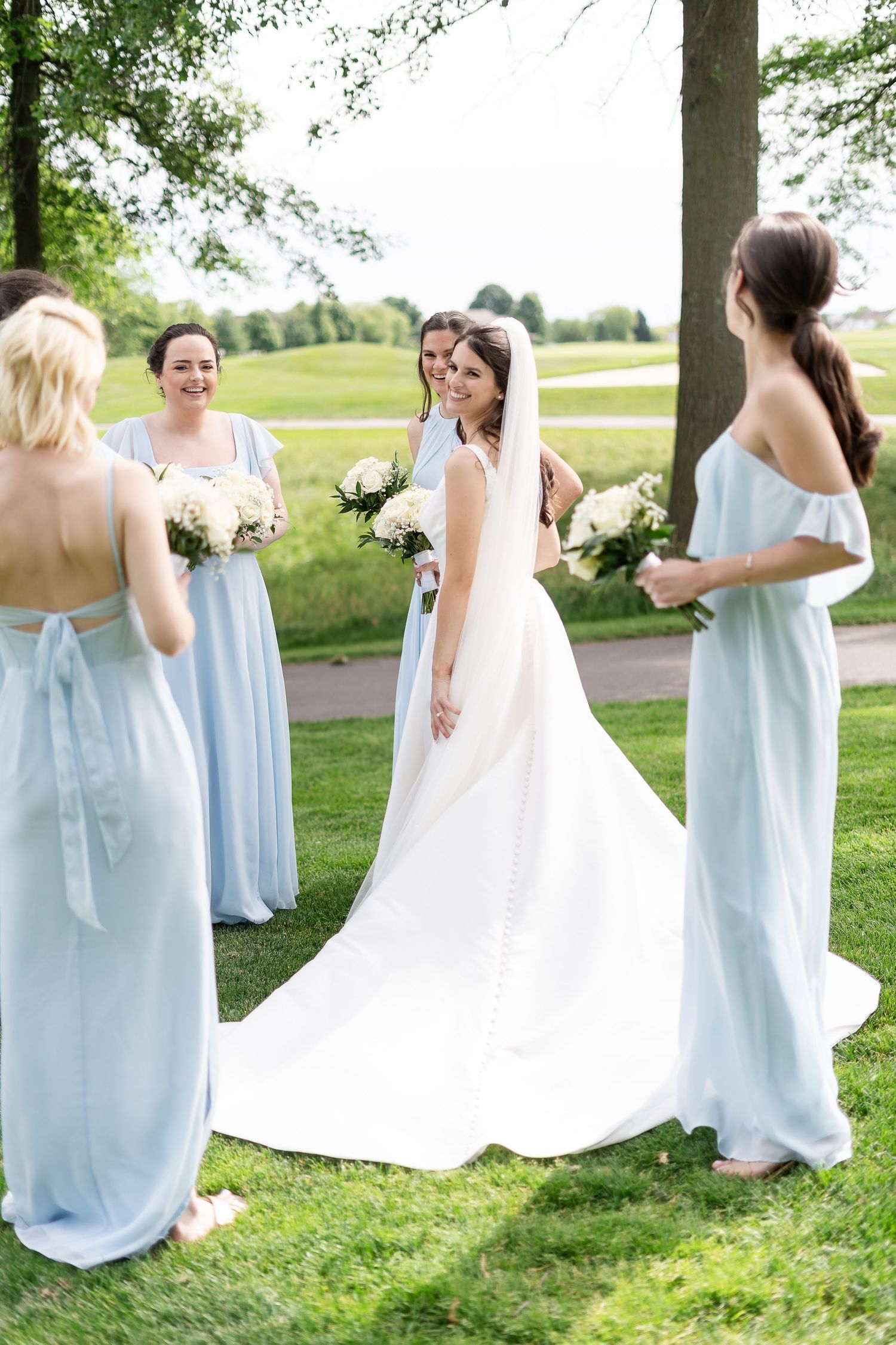 Golf course country club wedding. Bride surrounded by bridesmaids in light blue dresses holding white bouquets outdoors.