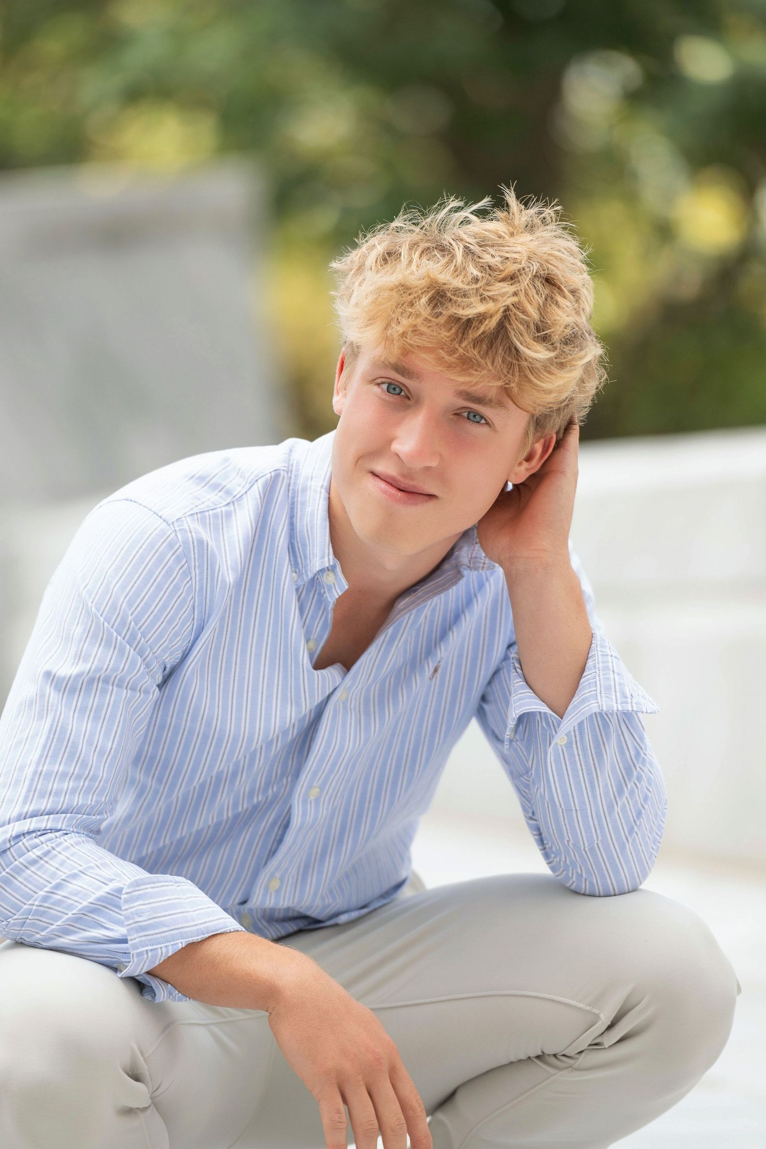 A person in a light blue striped button-down shirt and khaki pants poses casually on a white surface outdoors.