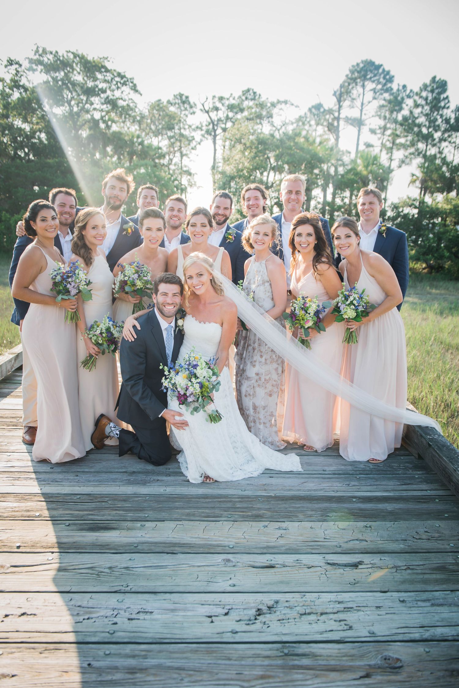 Wedding party poses together on rustic wooden boardwalk during outdoor summer celebration.