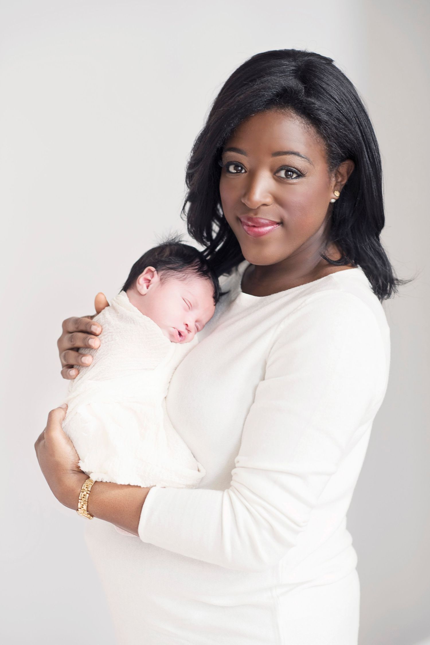 A woman in a white dress holds a sleeping newborn baby against her chest in a soft studio portrait.