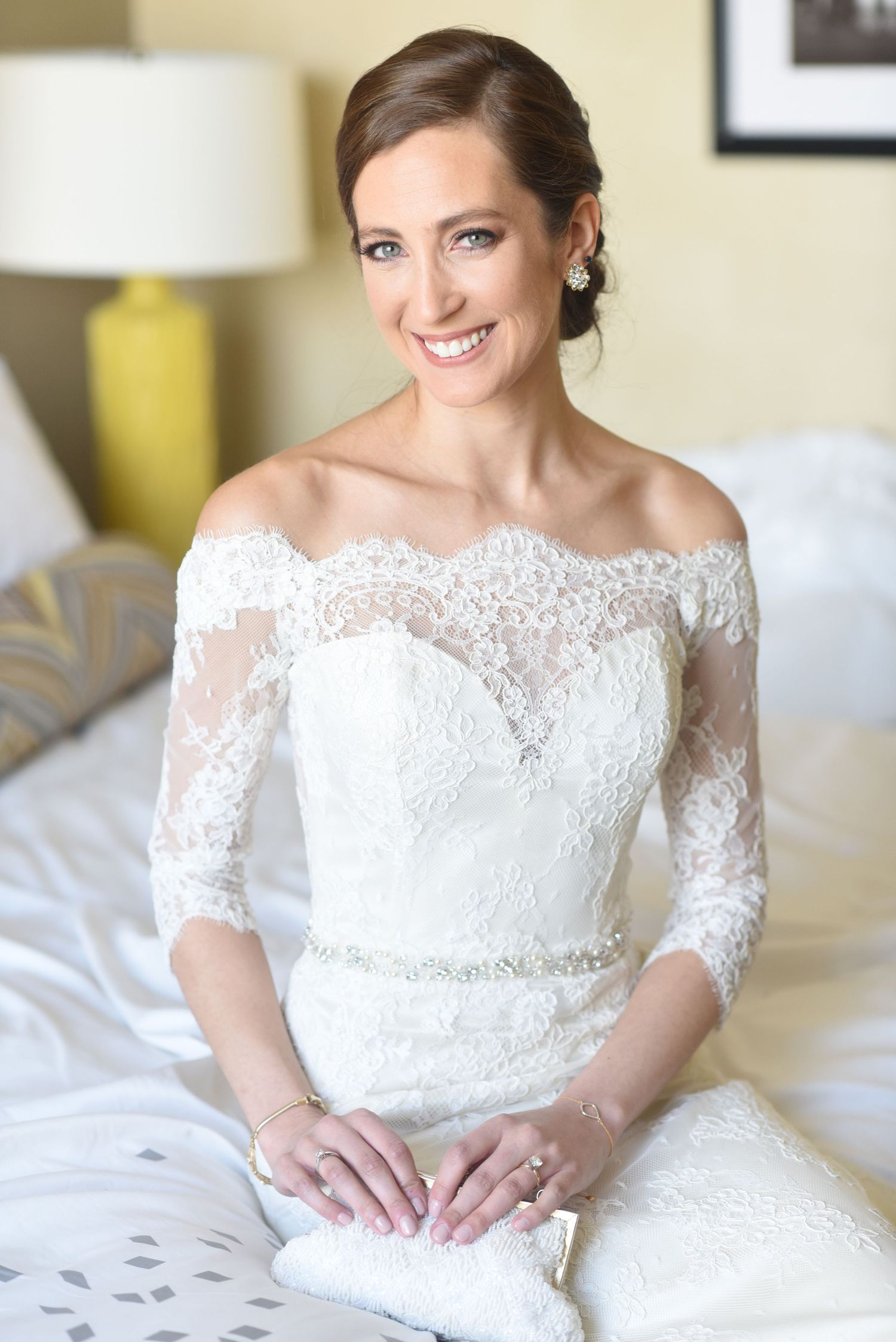 A bride in an off-shoulder lace wedding dress with three-quarter sleeves sits on a bed.