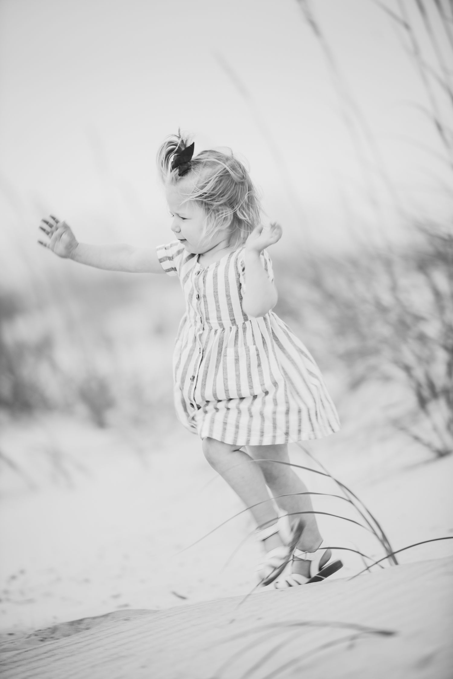 Black and white photo of a young girl in a striped dress playing on sandy beach dunes with sea grass.