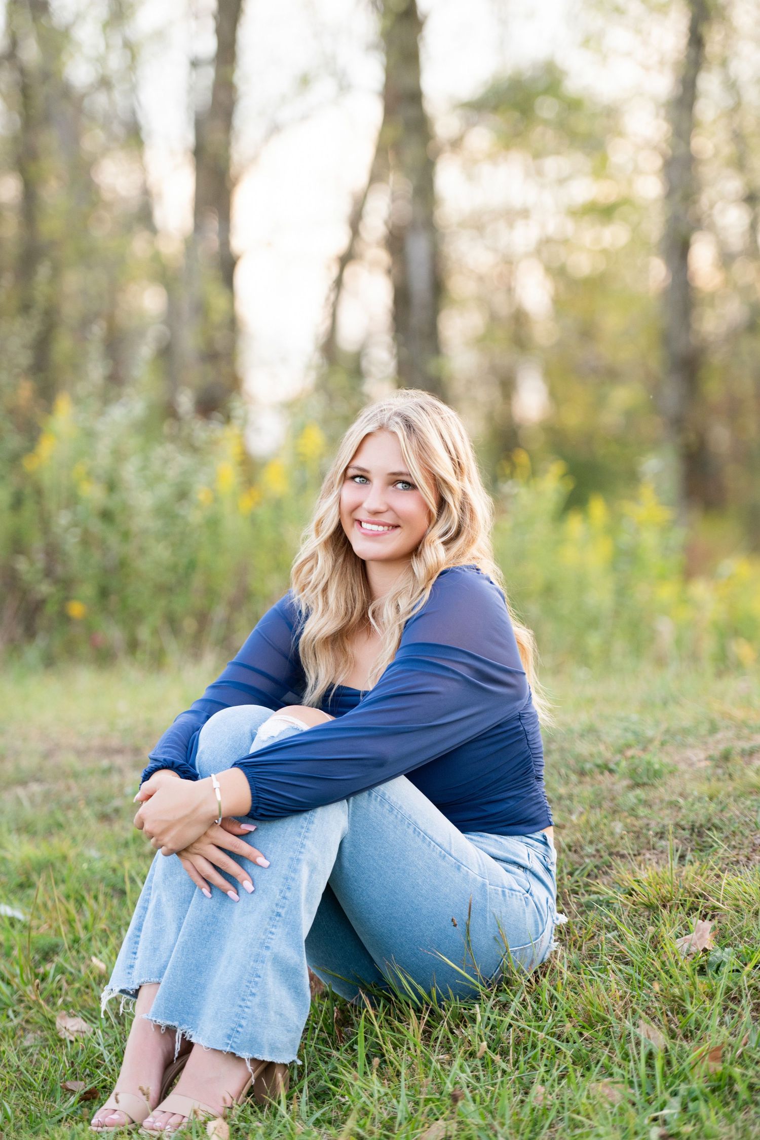 A Senior session in casual denim and navy blue top sits in grassy outdoor setting during golden hour. Alum Creek session