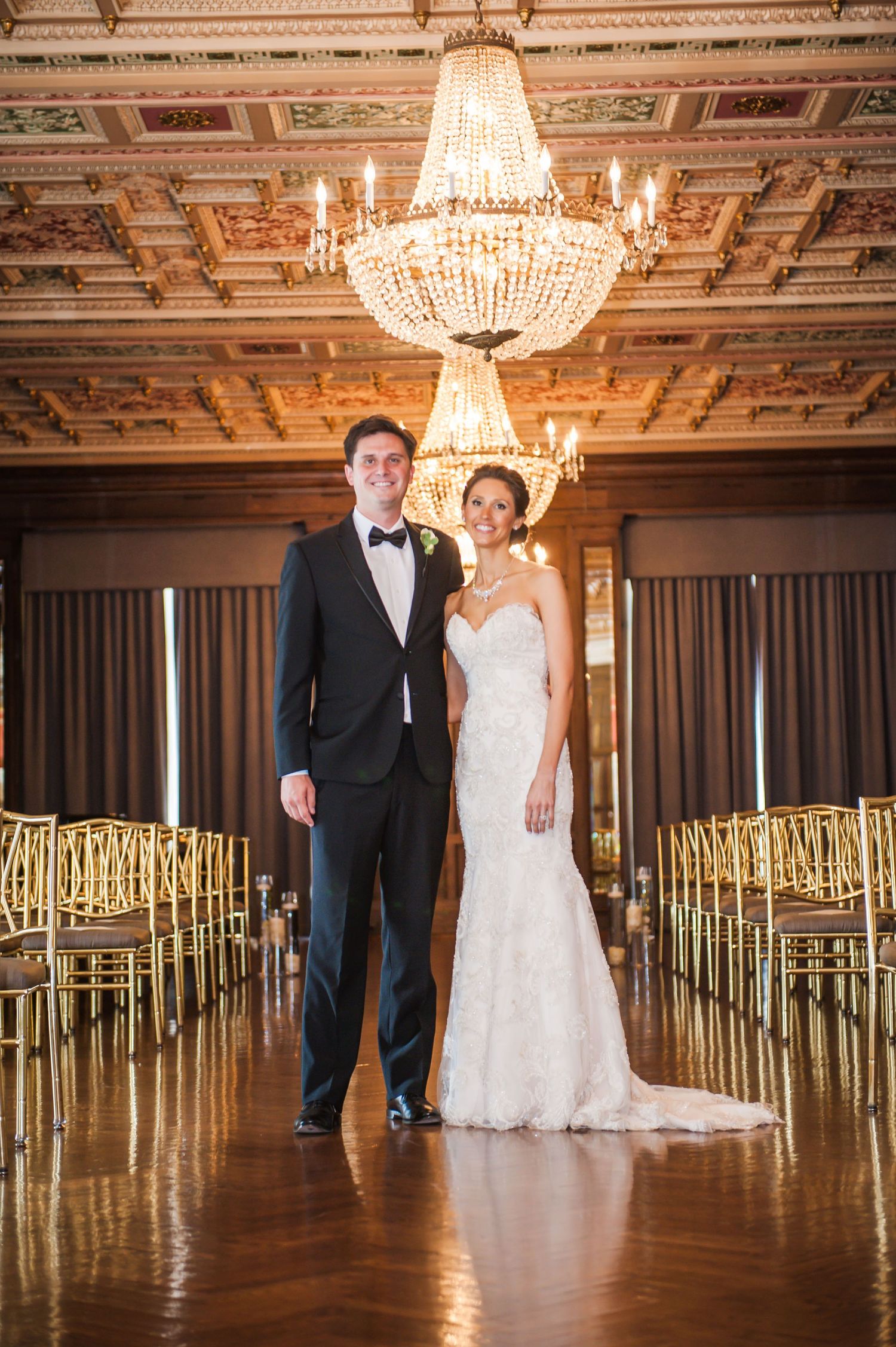 Elegant ballroom wedding venue with crystal chandeliers, ornate ceiling details, and gold chairs arranged in rows.
