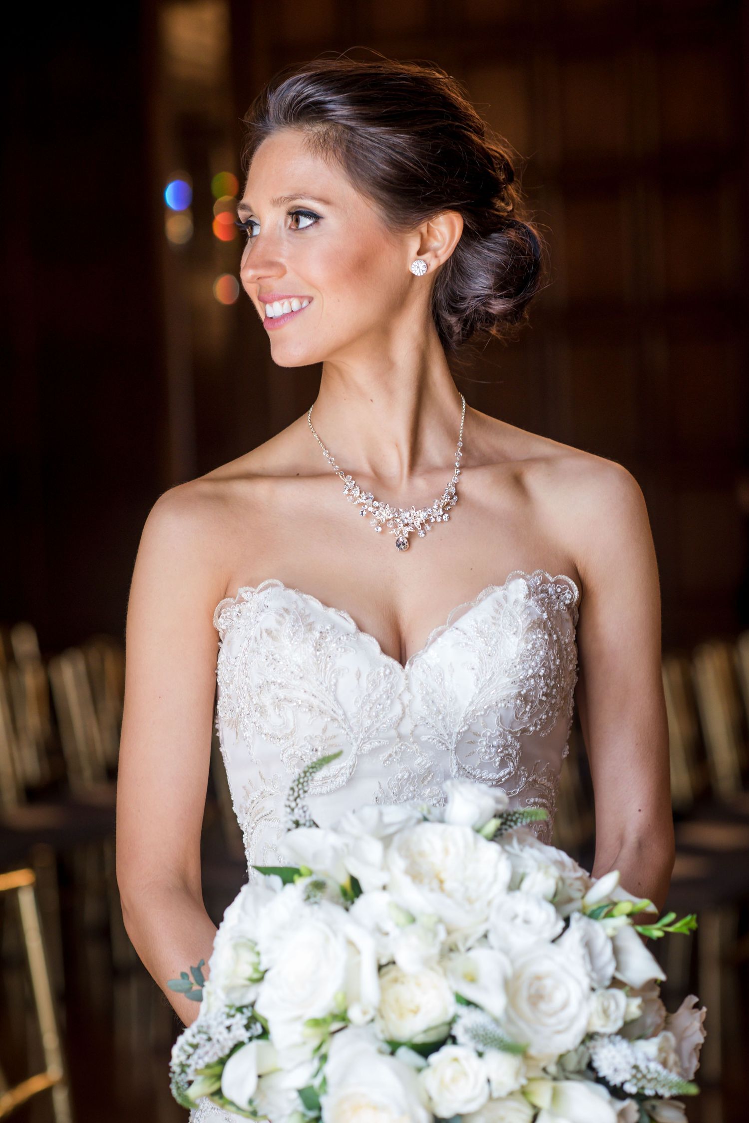 Bride in strapless lace wedding gown holds white rose bouquet while wearing elegant crystal necklace.
