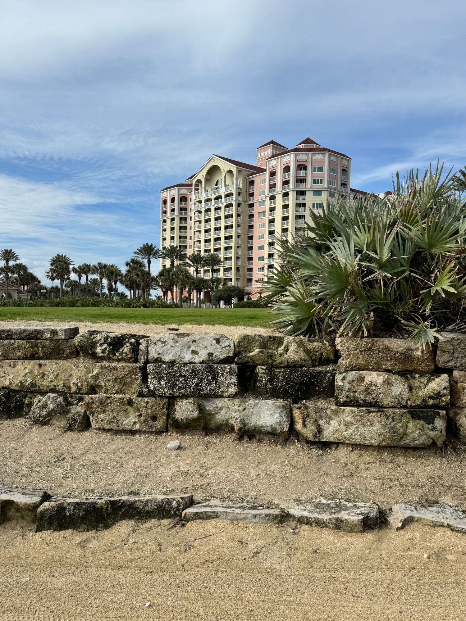Sandy beach with stone wall, palm trees, and a tall pink resort hotel under a partly cloudy blue sky.