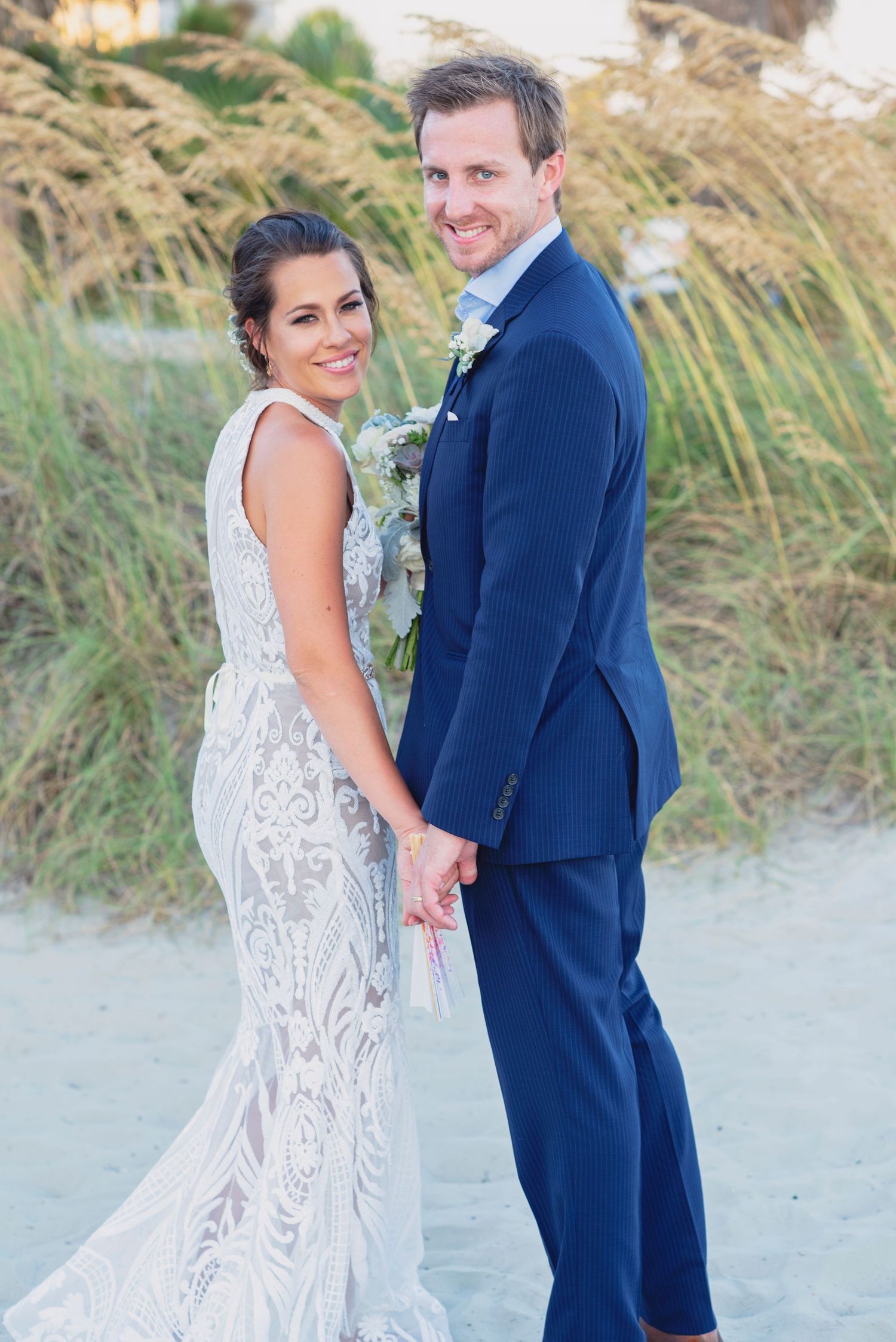 Bride and groom hold hands on a sandy beach, she in a lace gown, he in a navy suit with boutonniere.