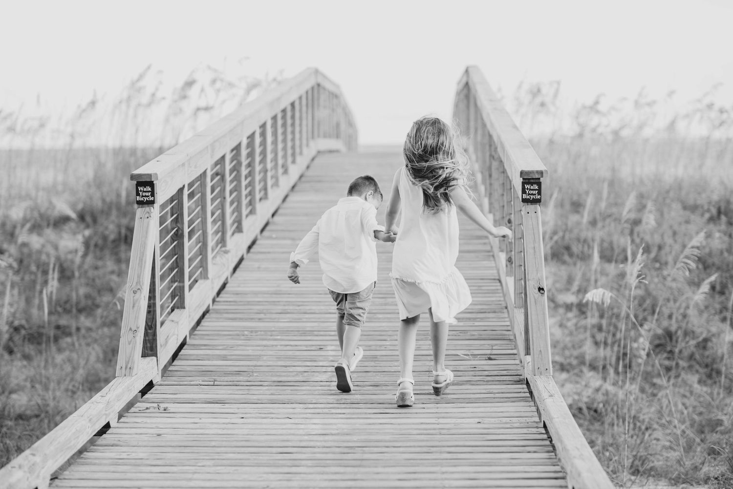 Two people walk hand in hand along a wooden boardwalk bridge through a grassy marsh area in black and white.